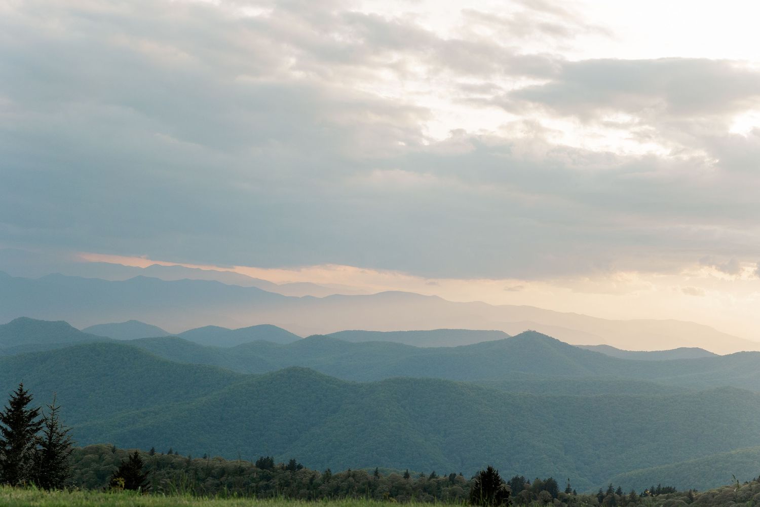 A peaceful mountain landscape at sunset with layered blue ridges stretching to the horizon under cloudy skies.
