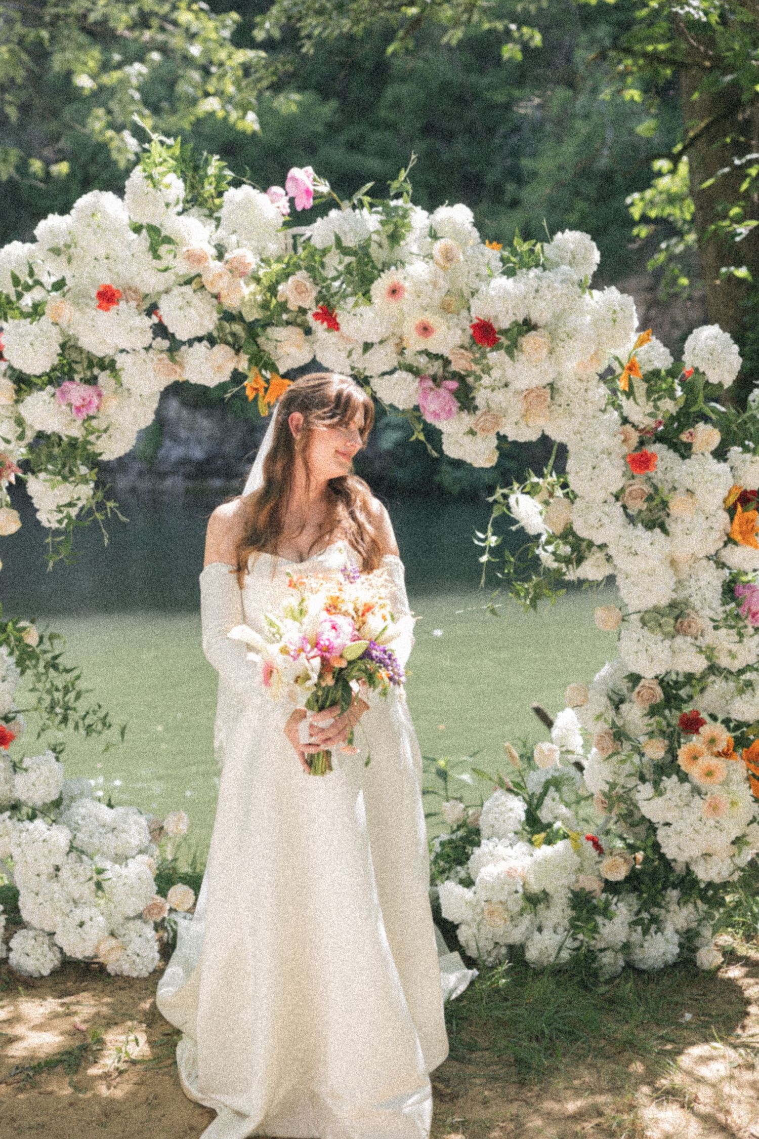 Bride stands under stunning white floral arch holding colorful bouquet by a serene river.