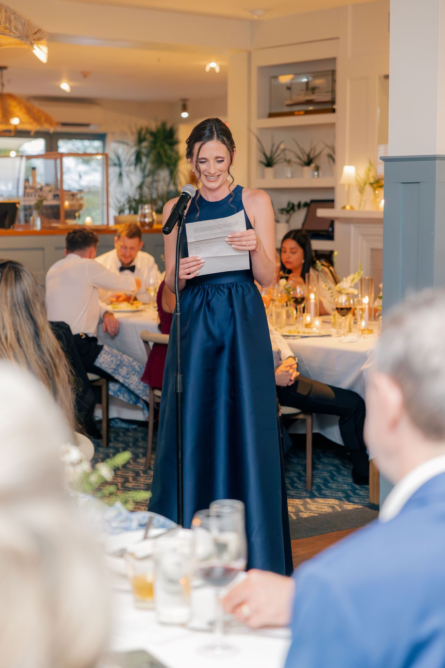 Bridesmaid in navy blue gown delivers a heartfelt speech at an elegant wedding reception dinner.