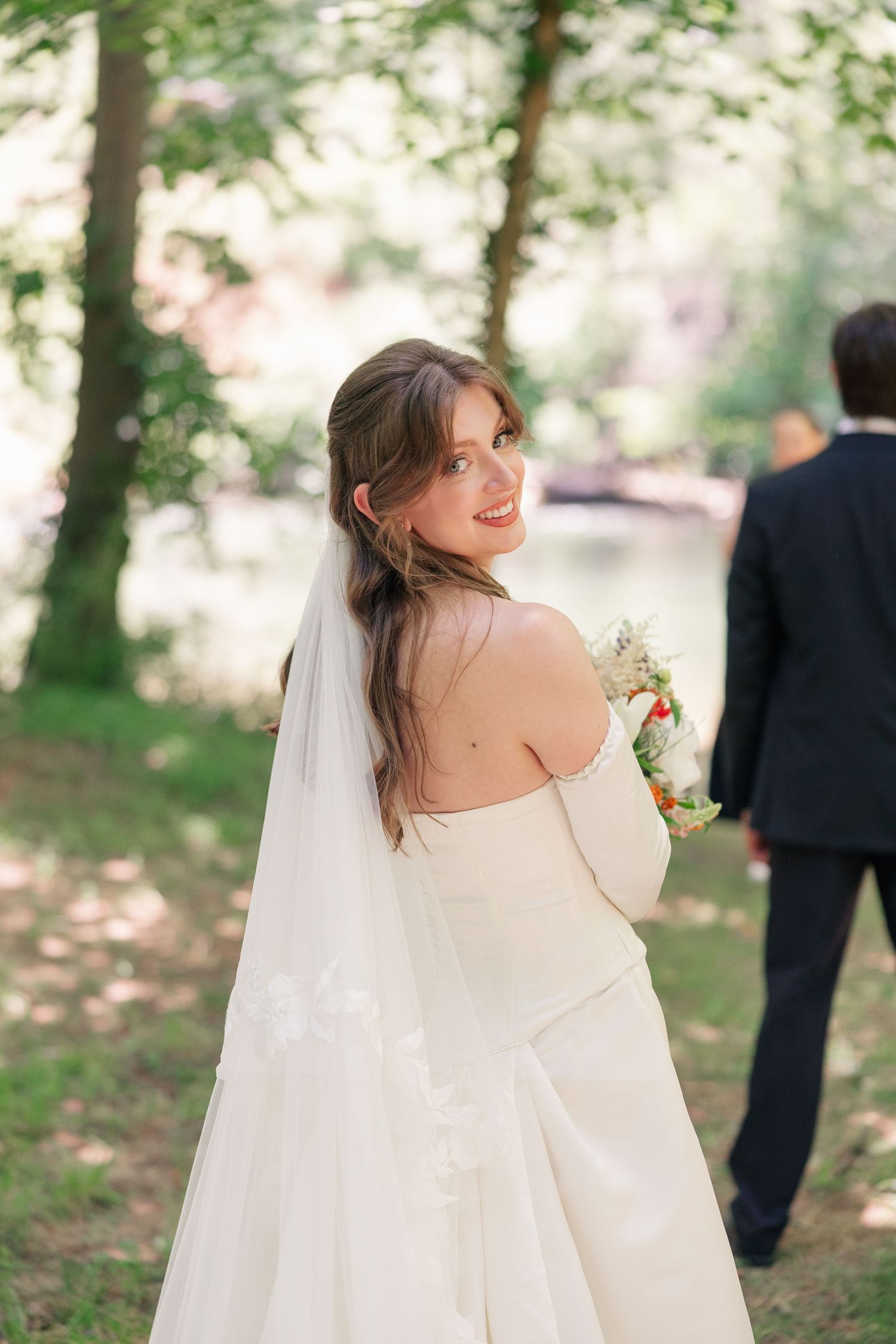 Bride in elegant white gown glances back with a smile holding a small floral bouquet.