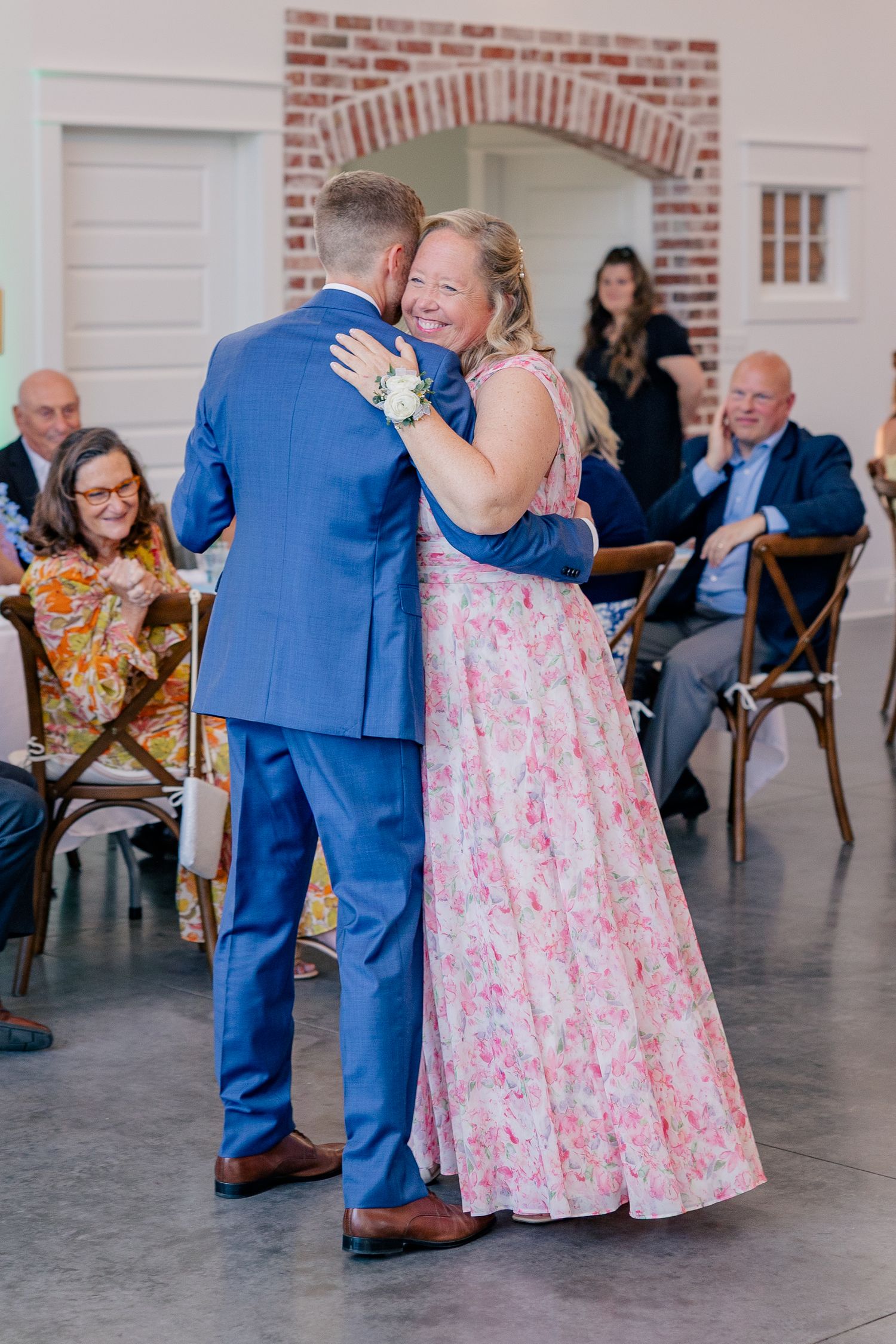 Groom in blue suit shares a warm dance with his mother in a pink floral dress at wedding reception.