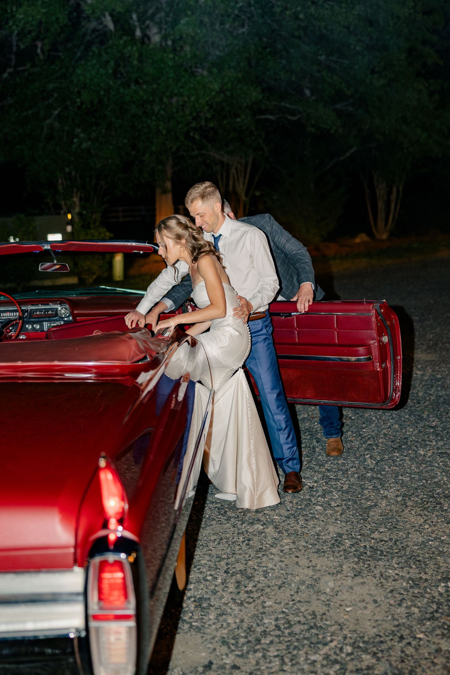 Bride and groom lean over vintage red classic car together at night after wedding reception.