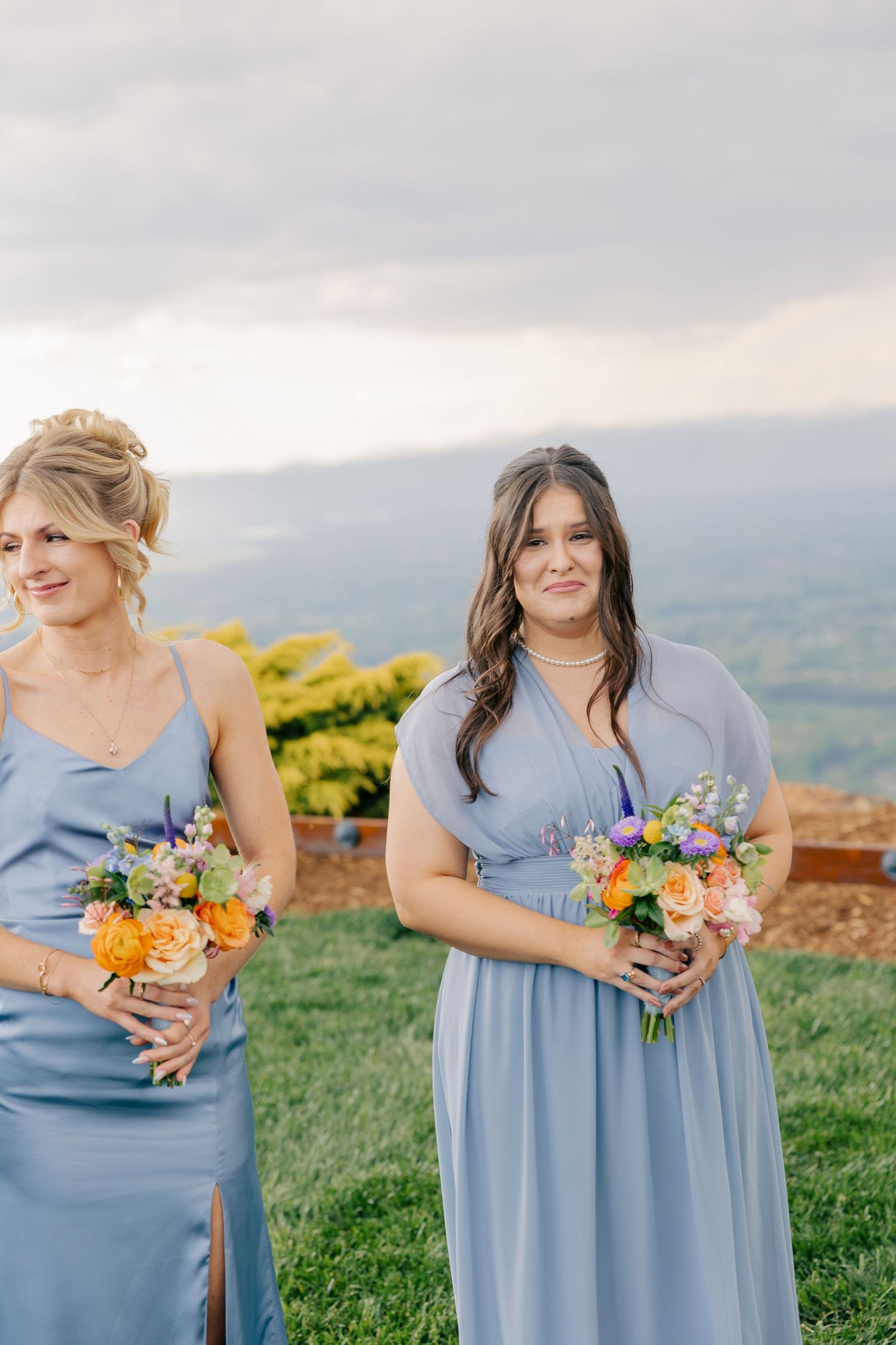 Two bridesmaids in dusty blue gowns holding colorful bouquets at outdoor hilltop wedding ceremony.