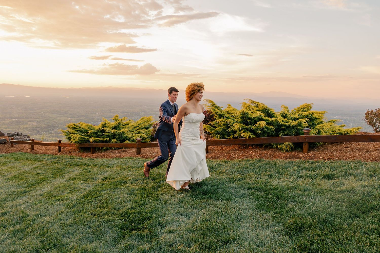Bride and groom run across green lawn at sunset with panoramic mountain valley views behind them.