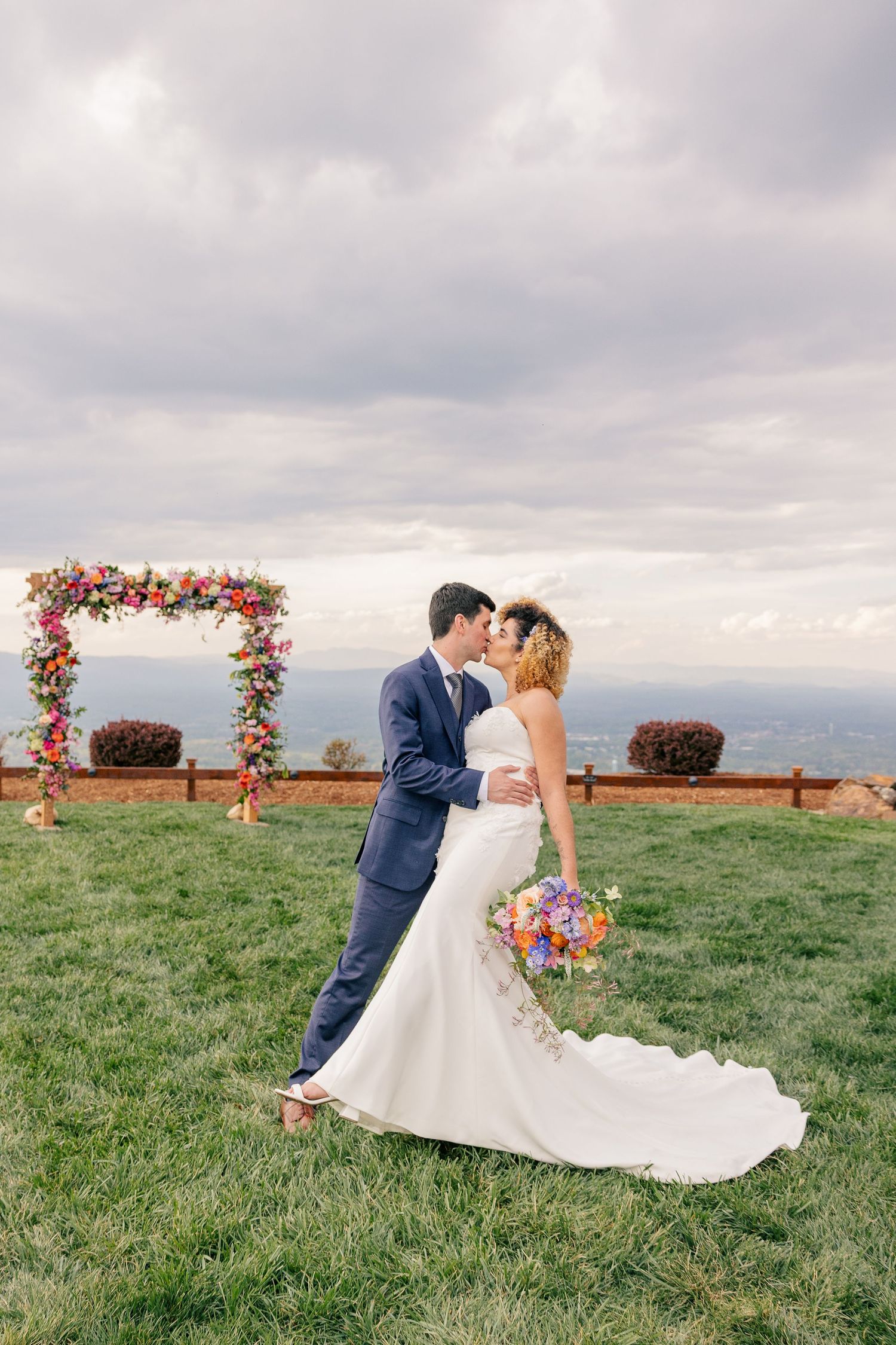 Bride and groom share a kiss outdoors near a colorful floral arch with mountain views on their wedding day.