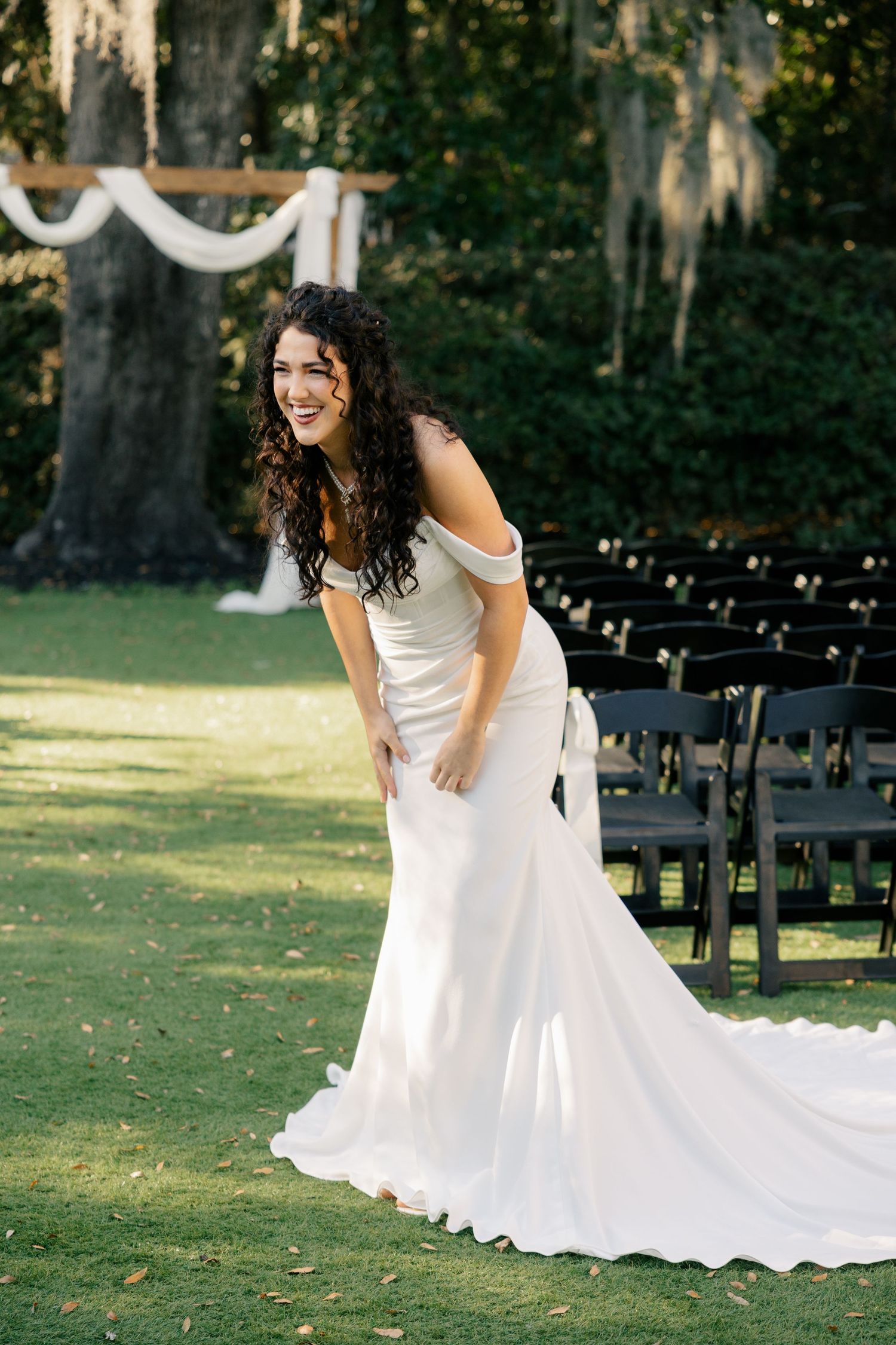 A beautiful wedding dress with a flowing train photographed outdoors on a lawn with decorative chairs and hanging drapery.