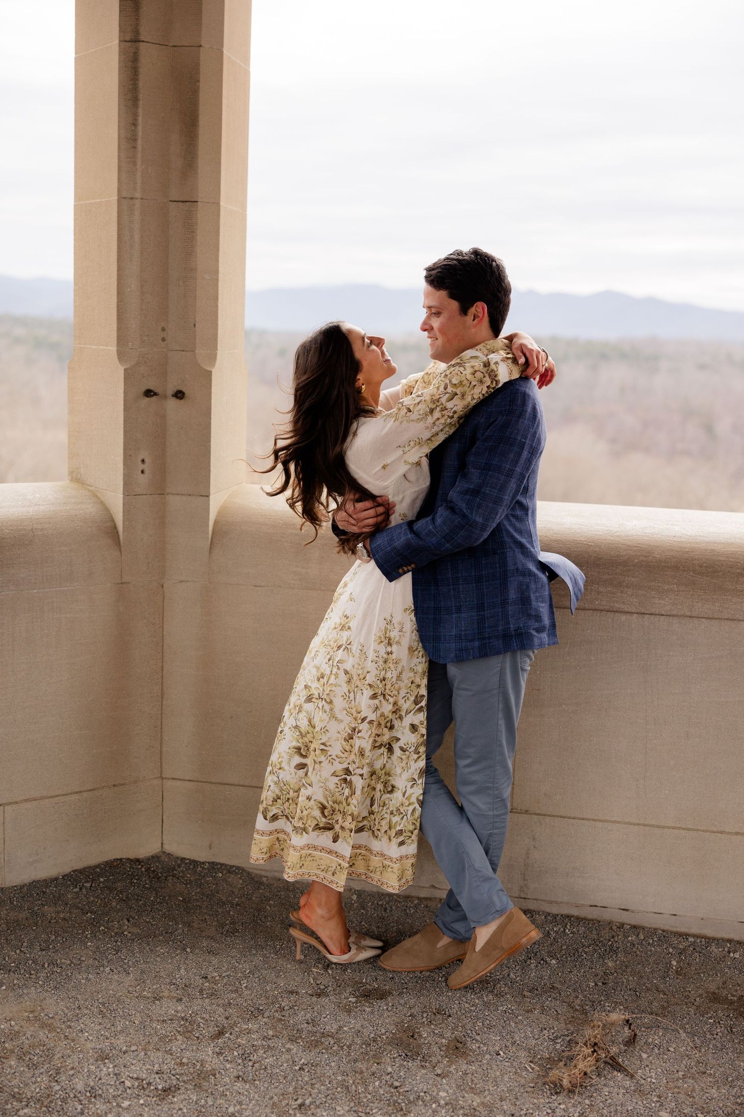 A romantic couple embraces on a balcony overlooking mountain views, wearing stylish clothing for an engagement photoshoot.