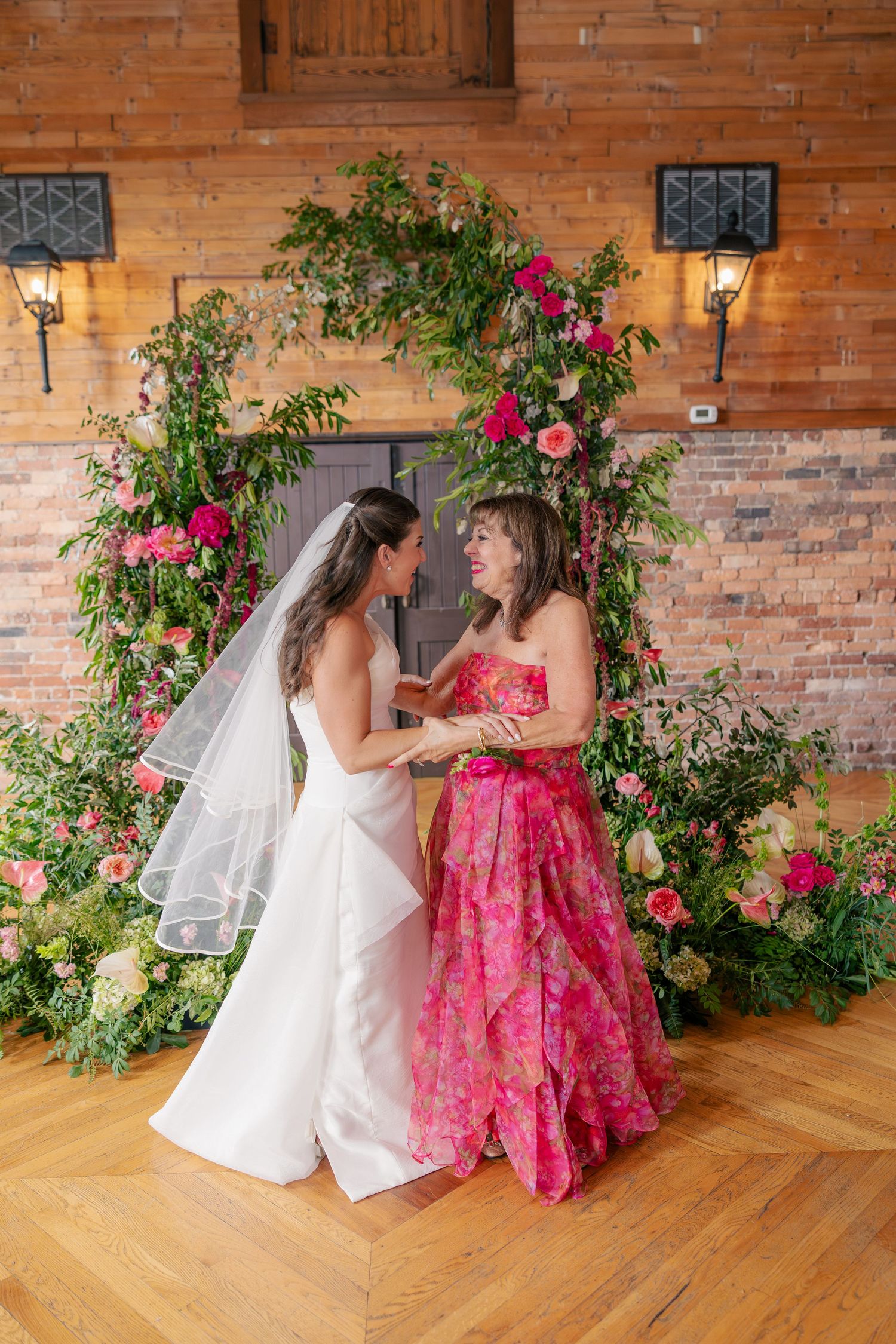 Bride and woman in pink floral gown hold hands before lush floral arch at wedding ceremony.