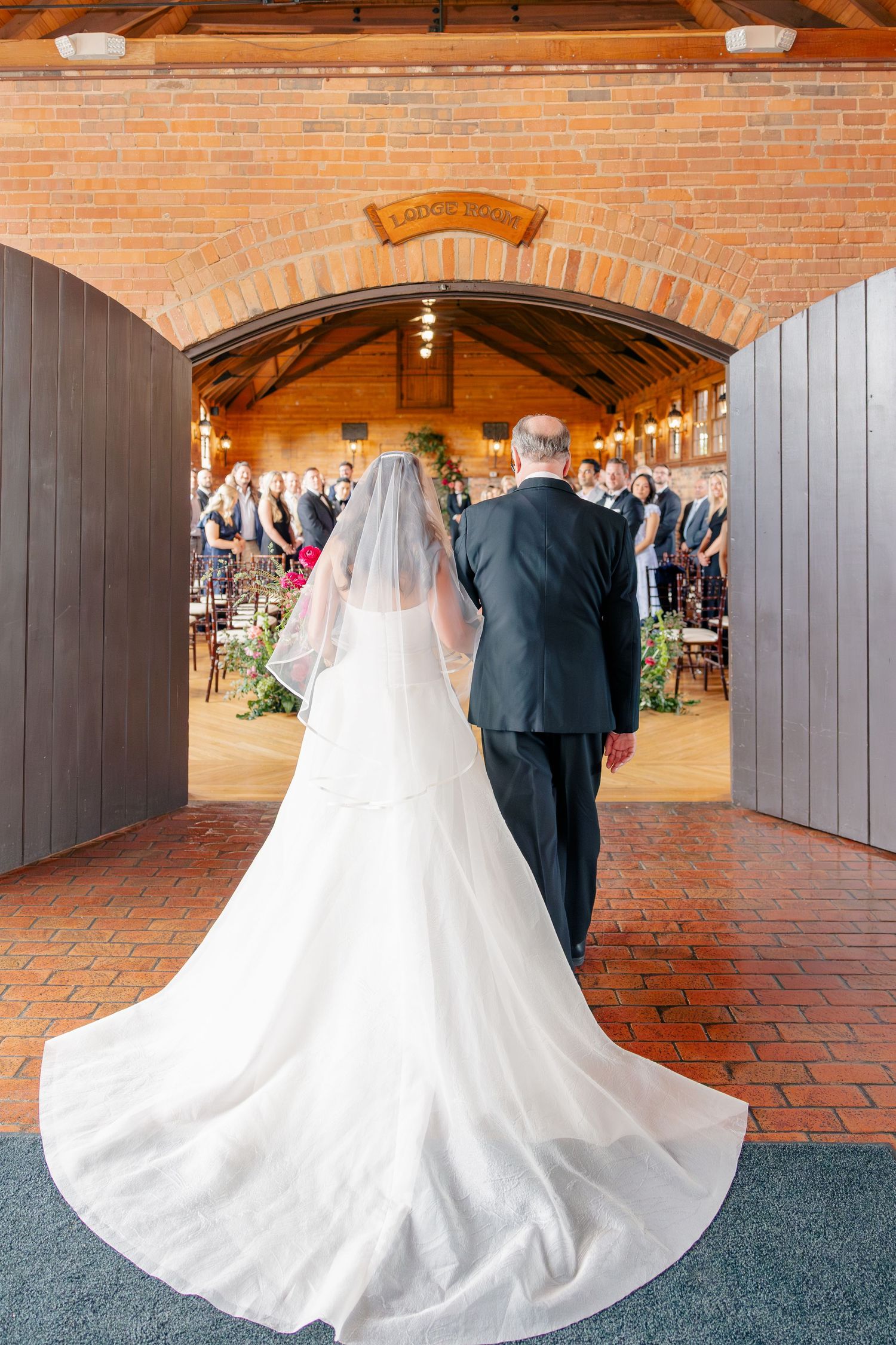 Bride in flowing white gown and veil walks down aisle with escort through brick archway toward wedding guests.
