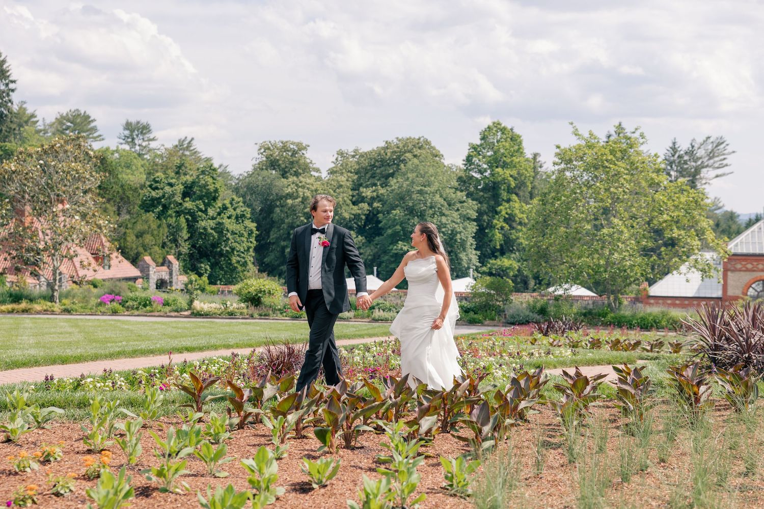 Bride and groom walk hand in hand through lush garden grounds at their outdoor summer wedding.