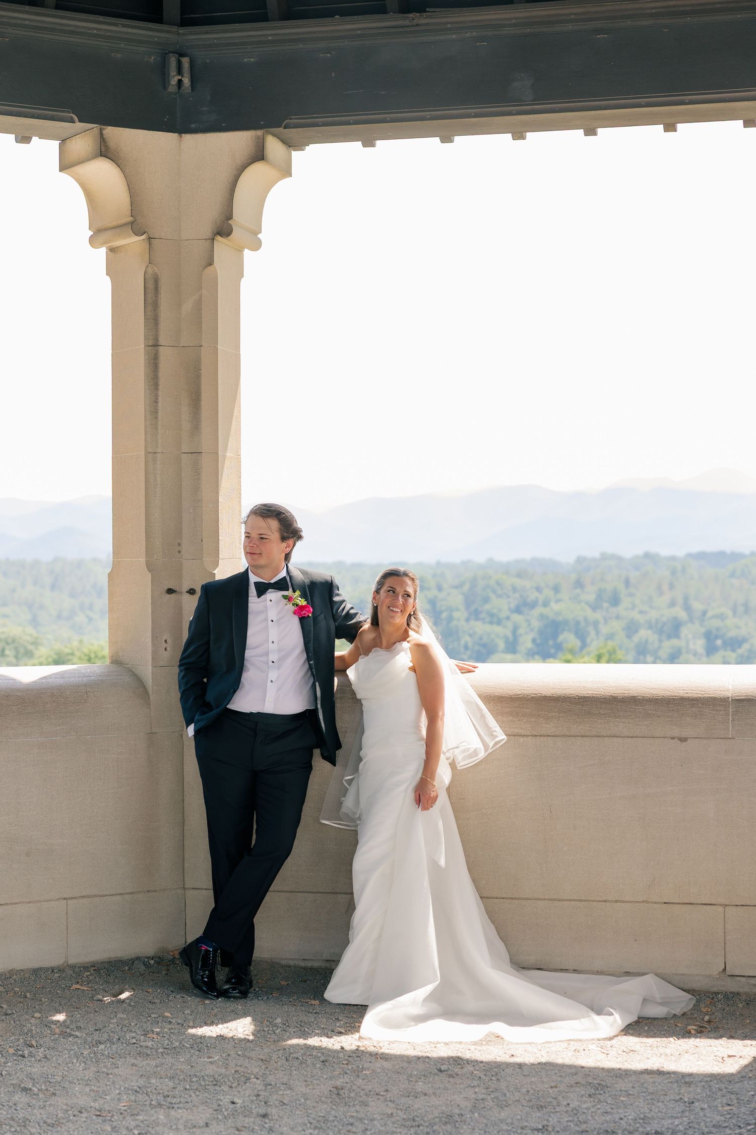 Bride and groom pose together in a sunlit stone pavilion with scenic mountain views in the background.