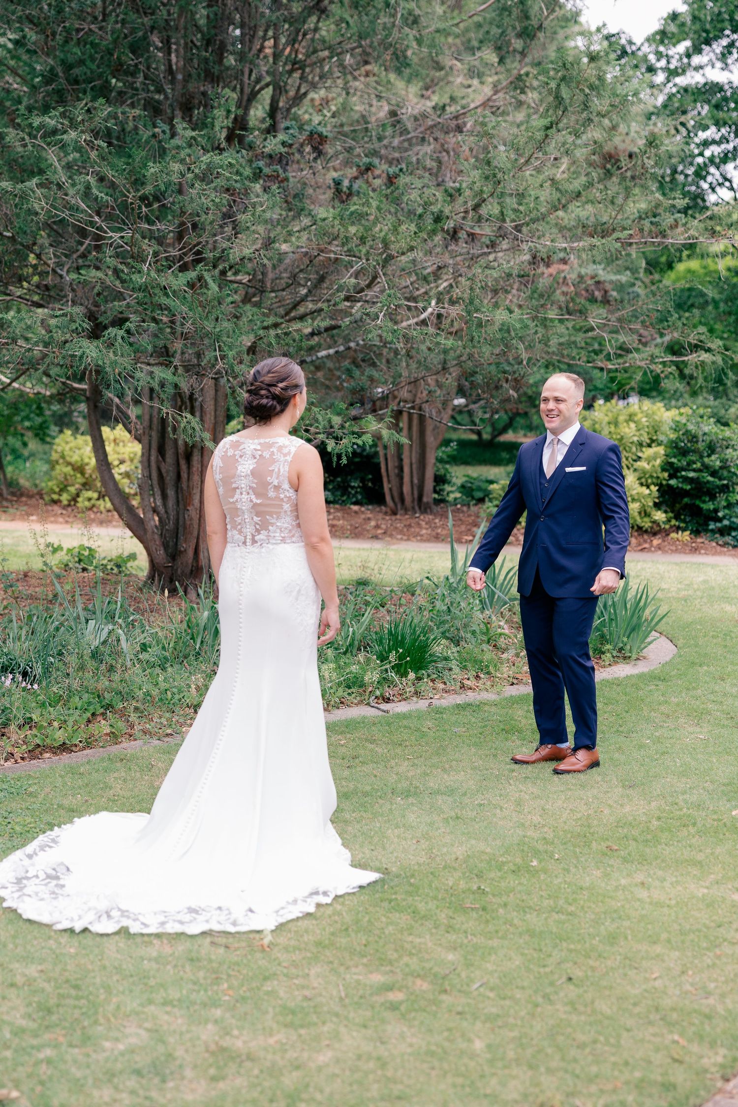 Bride in lace wedding gown shares first look moment with groom in navy suit on lush green lawn.