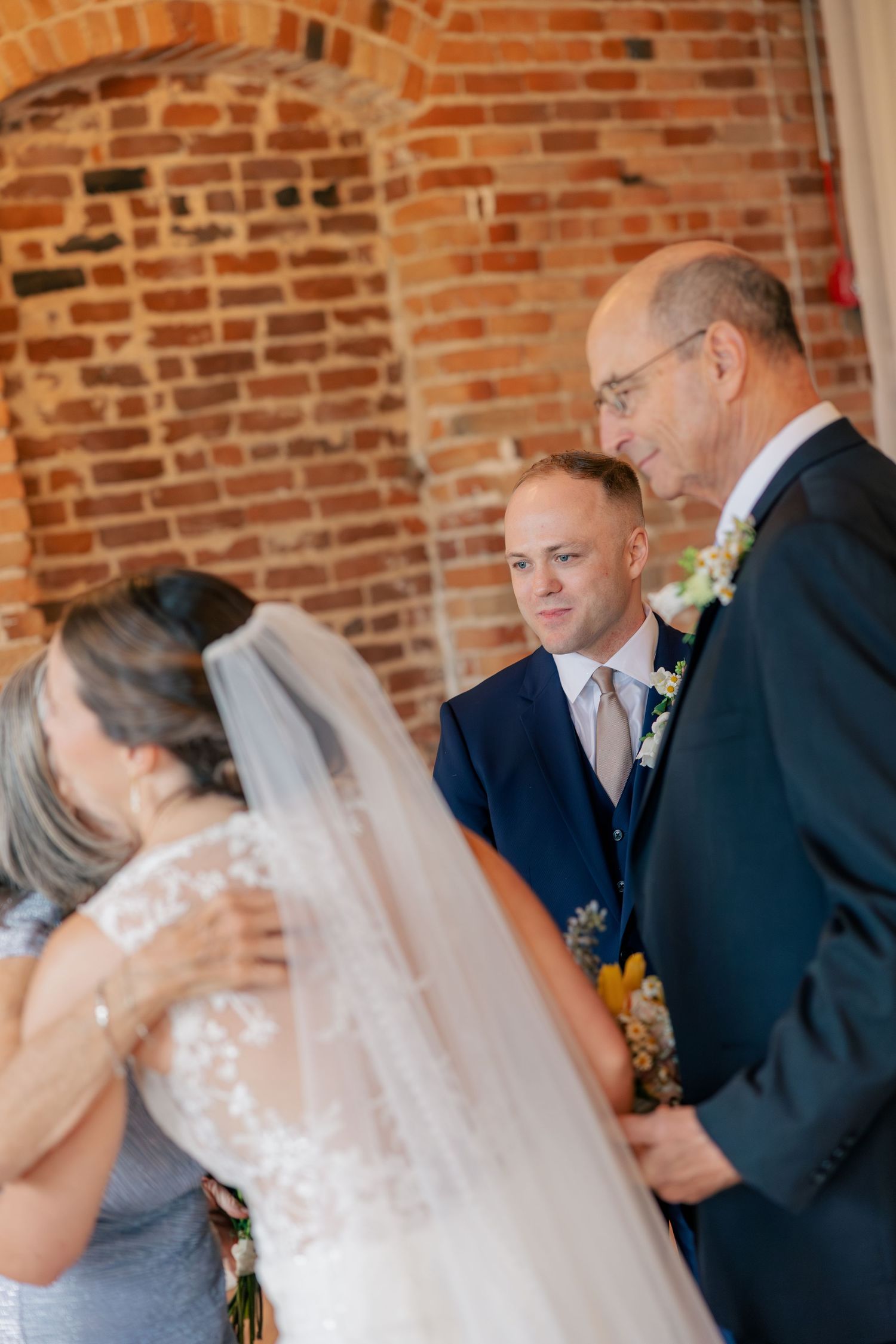 Groom watches emotionally as bride shares a tender moment with family before wedding ceremony in brick venue.