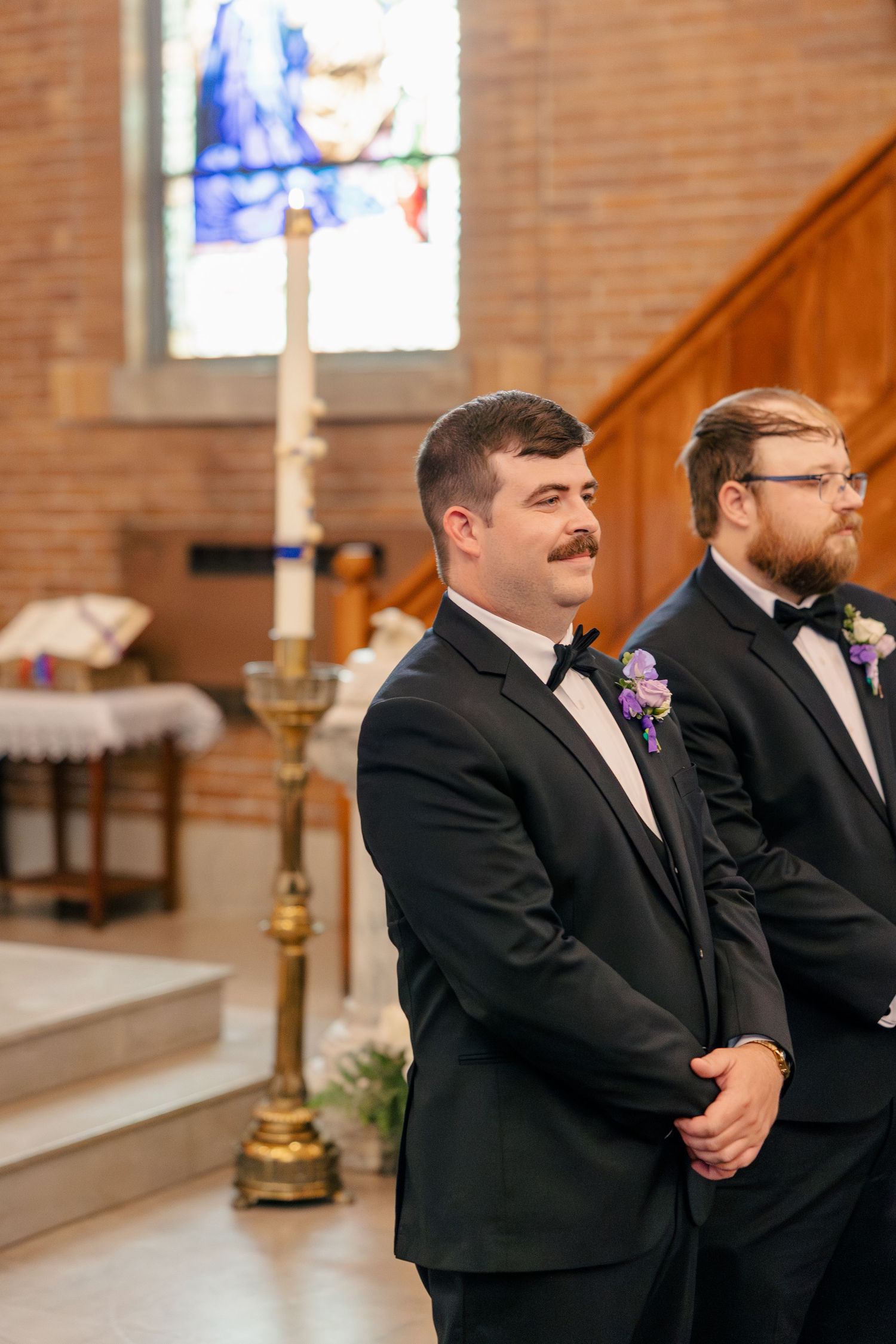 Groom in black tuxedo smiles at altar inside brick church with stained glass windows during ceremony.