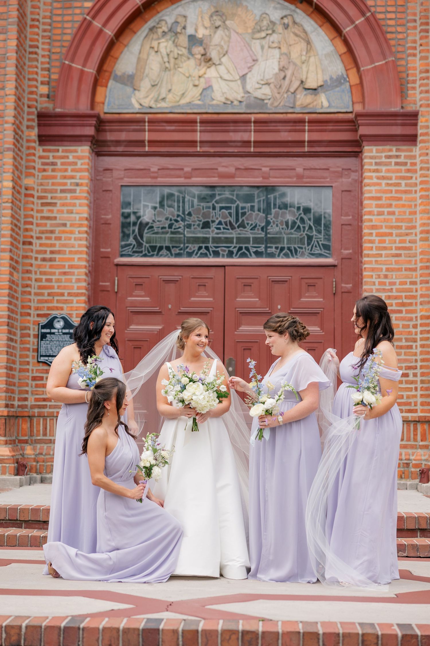 Bride with bridesmaids in lavender gowns outside ornate red brick church entrance.