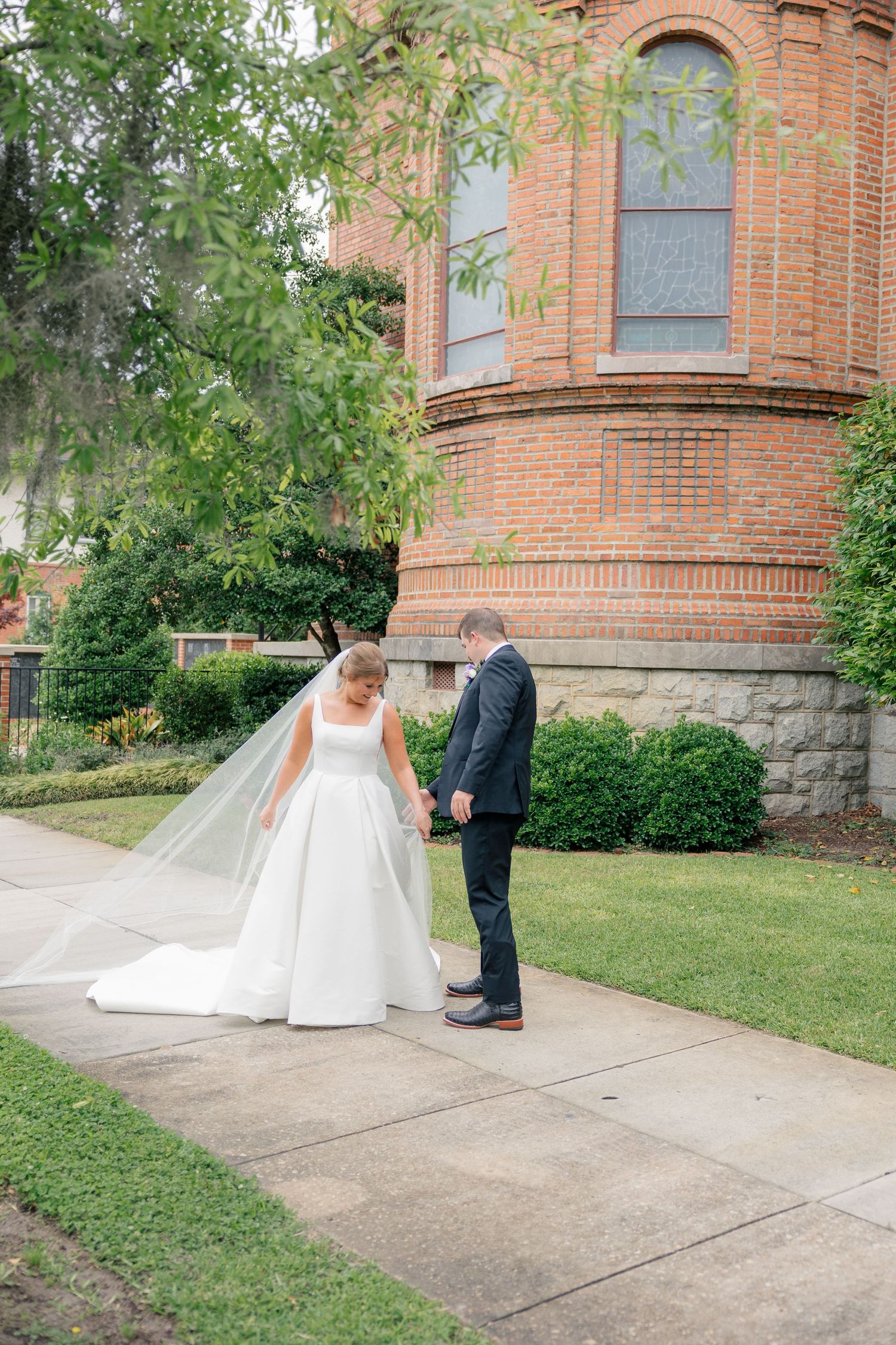 Bride and groom share first look moment outside historic red brick church surrounded by greenery.
