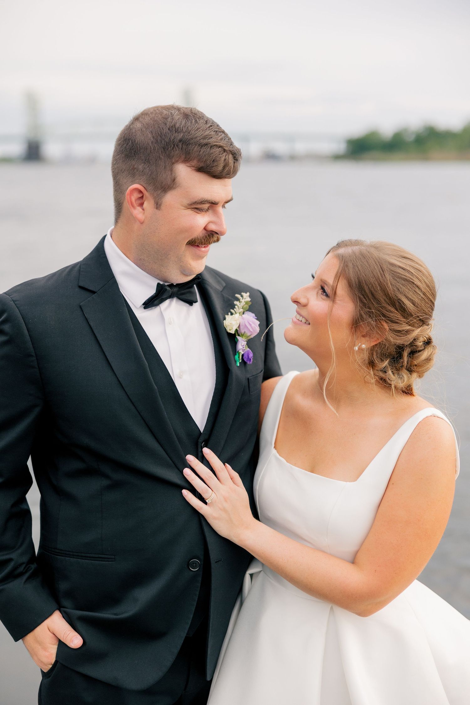 A bride and groom share a joyful moment near a waterfront, dressed in wedding attire with a boutonniere and white gown.