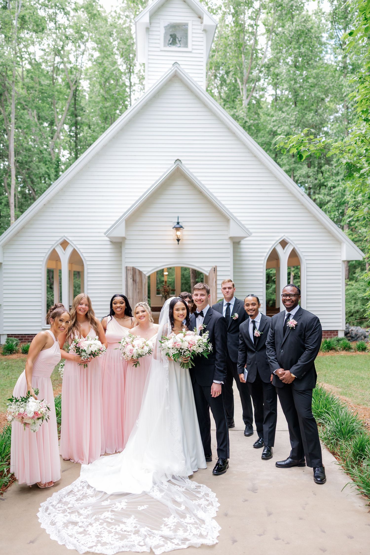 Wedding party poses in front of charming white chapel with pink bridesmaid dresses and formal attire.