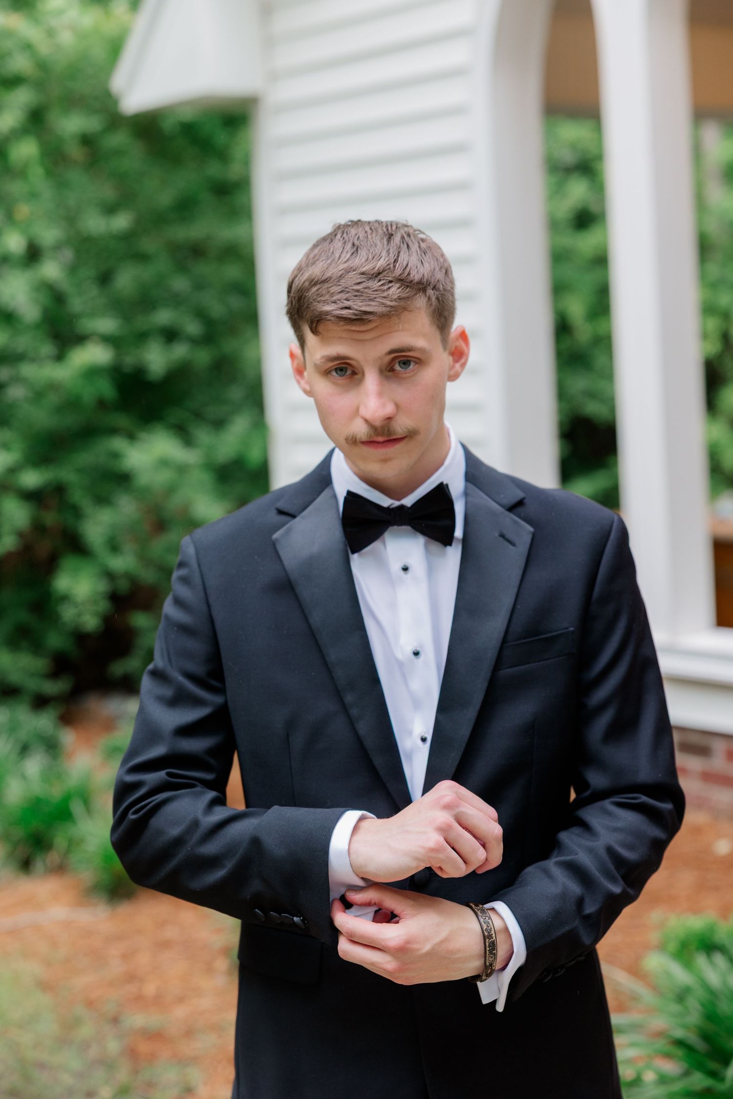 A person in a black tuxedo and bowtie adjusts their cufflinks outdoors.