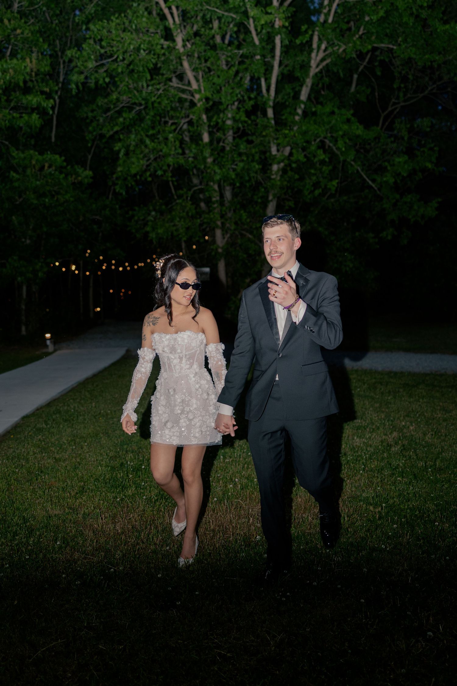A couple taking romantic nighttime photos in a wooded area illuminated by string lights.