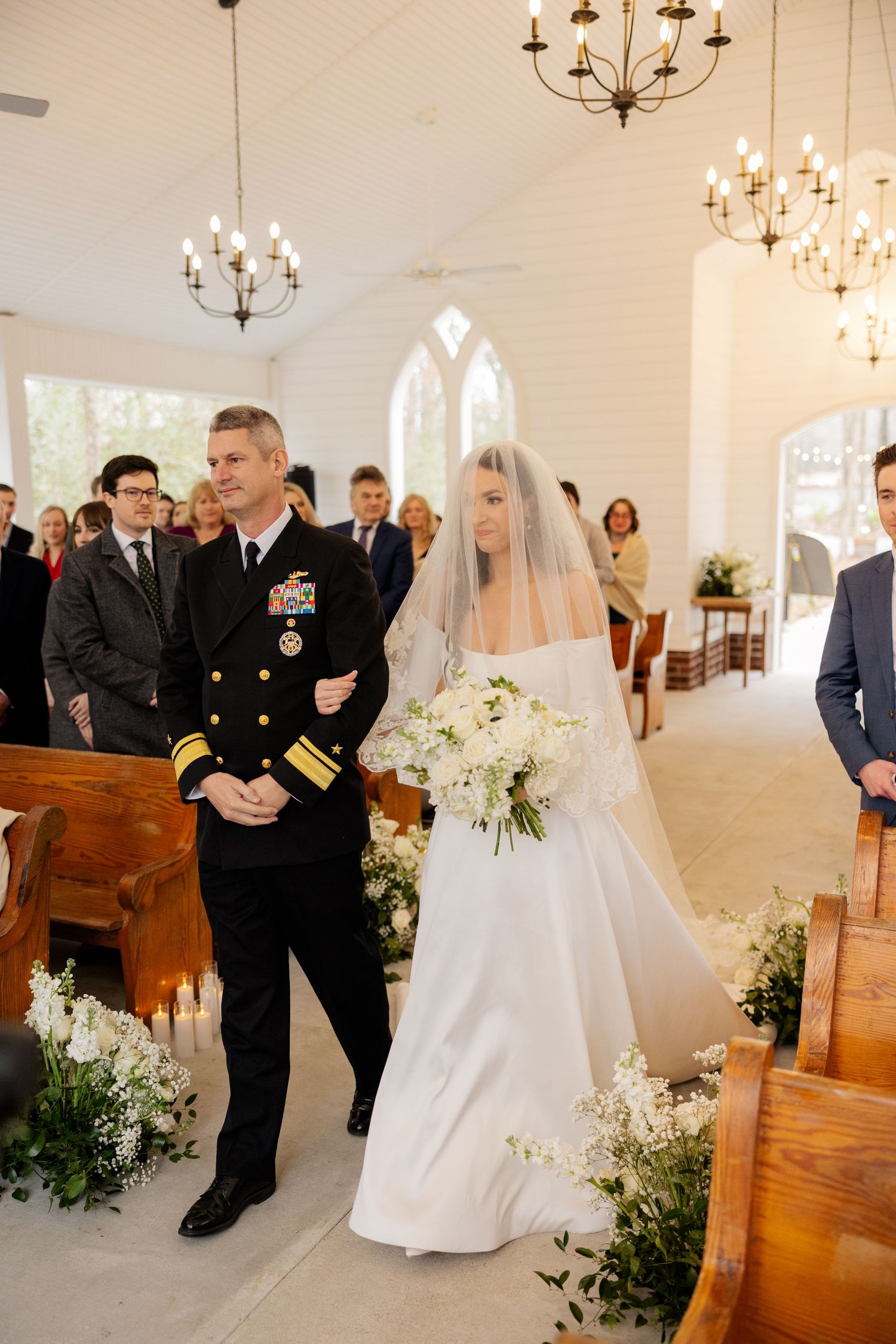 White chapel wedding ceremony with chandeliers and floral arrangements lining the aisle.