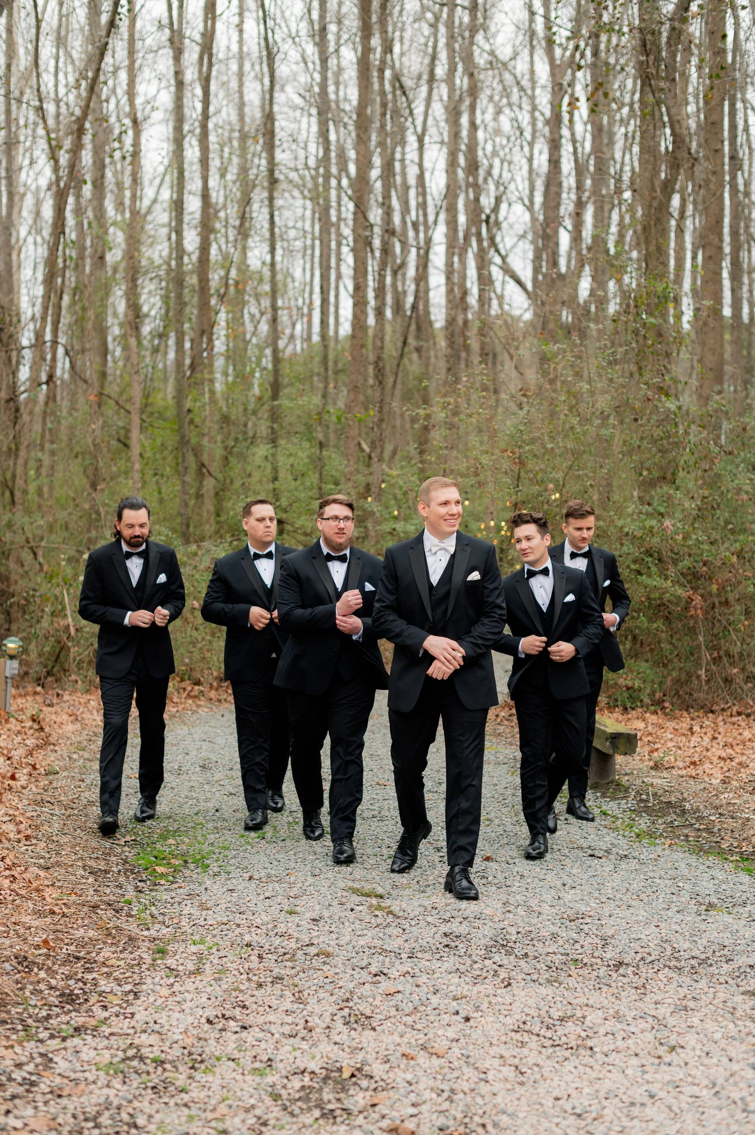 A group of groomsmen in black tuxedos walk along a gravel path surrounded by bare trees in winter.