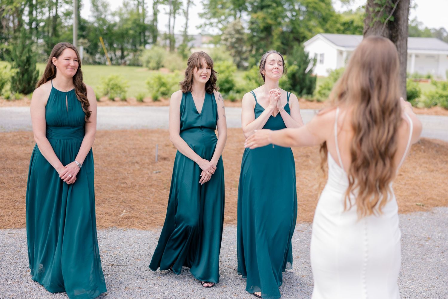 Bridesmaids in elegant teal green floor-length dresses stand on gravel path while looking at bride in white gown.