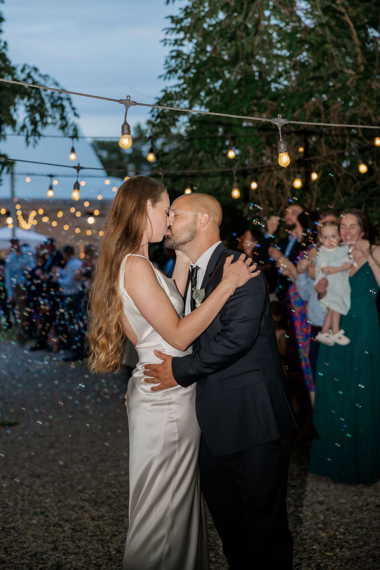 A romantic evening wedding dance under twinkling string lights with guests in the background.