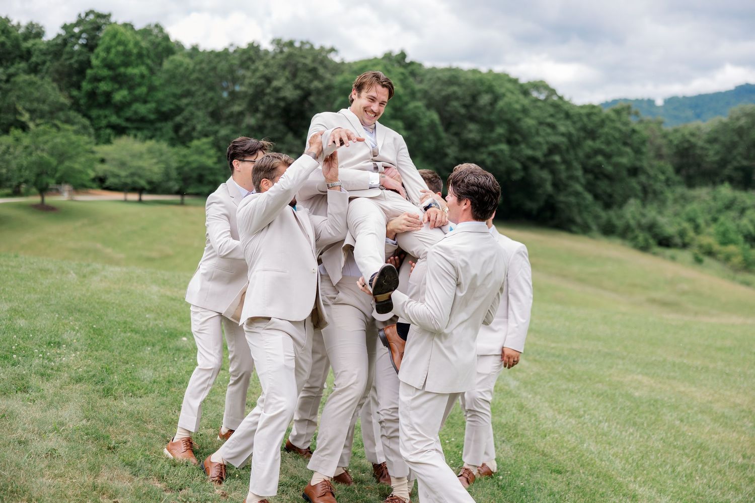 Groomsmen in light beige suits celebrate together on a rolling green hillside on a cloudy summer day.