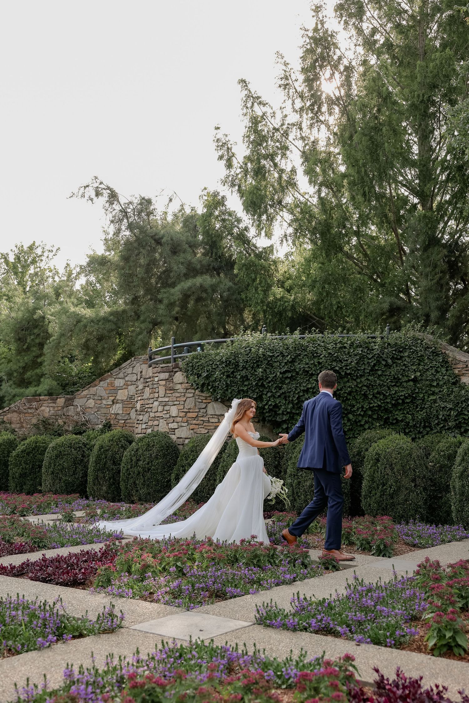 Couple walking through a garden path lined with purple flowers and stone walls.