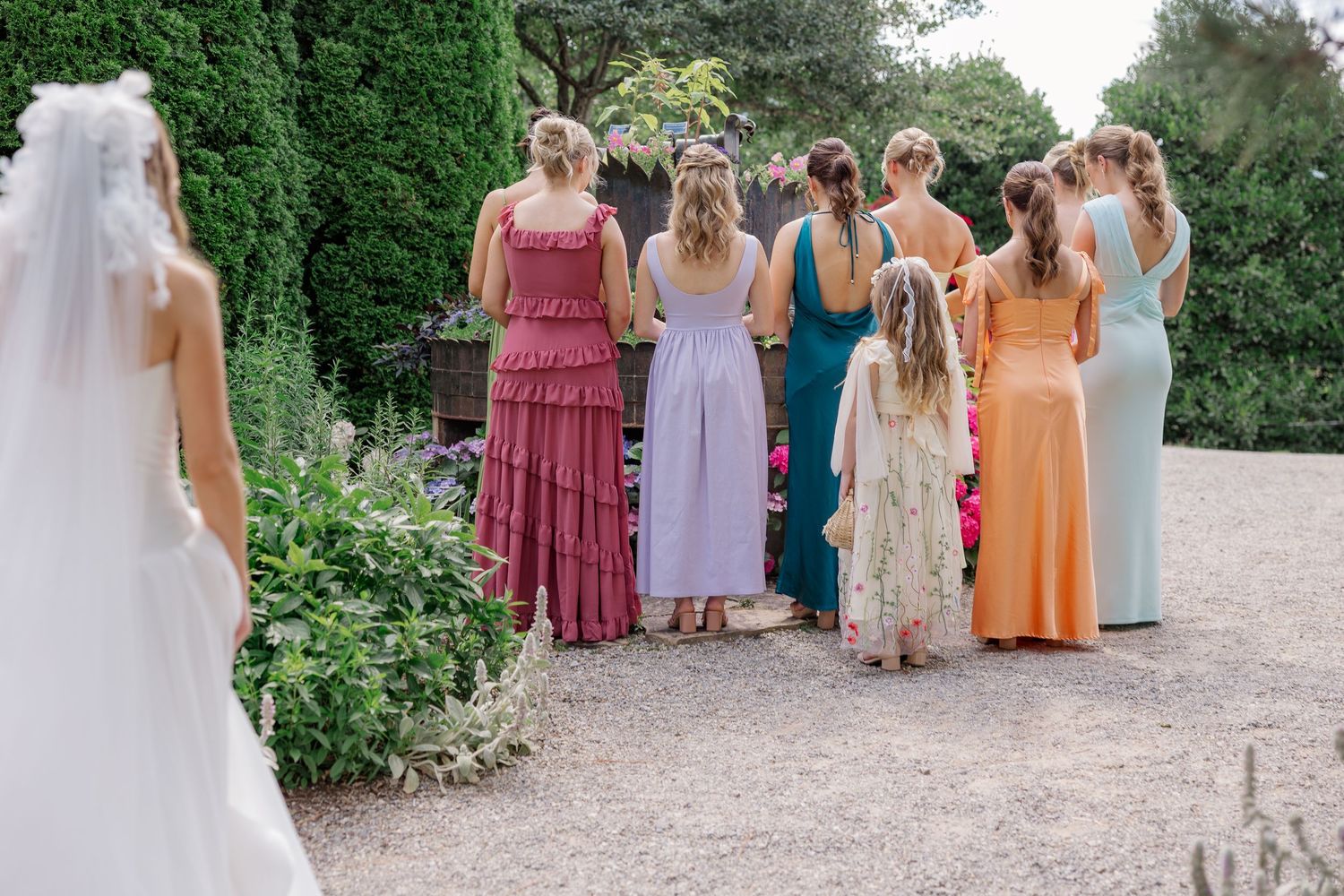 A wedding party of women in colorful dresses stands on a garden path lined with manicured hedges.