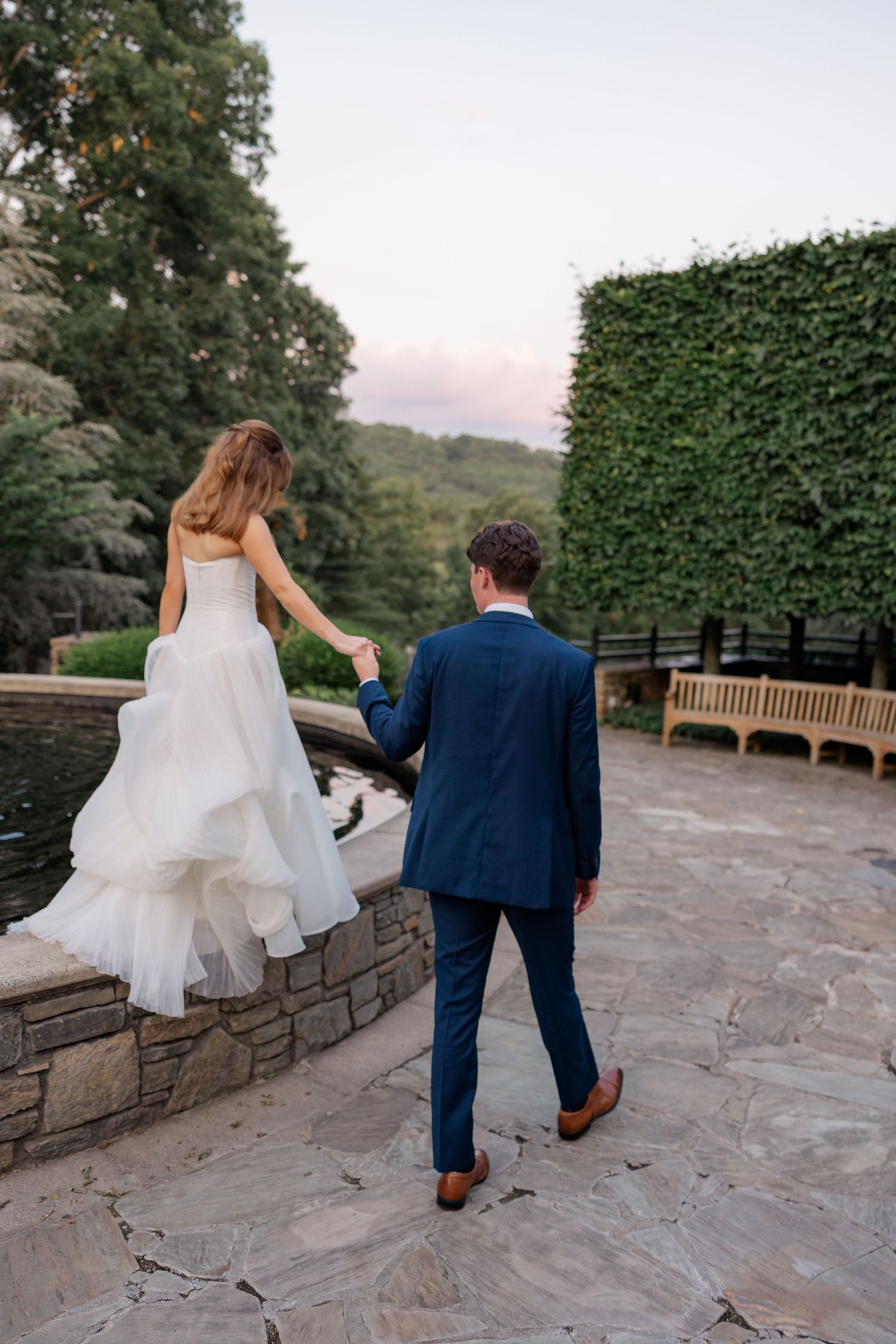 A couple walks along a stone wall path surrounded by lush greenery and garden benches.