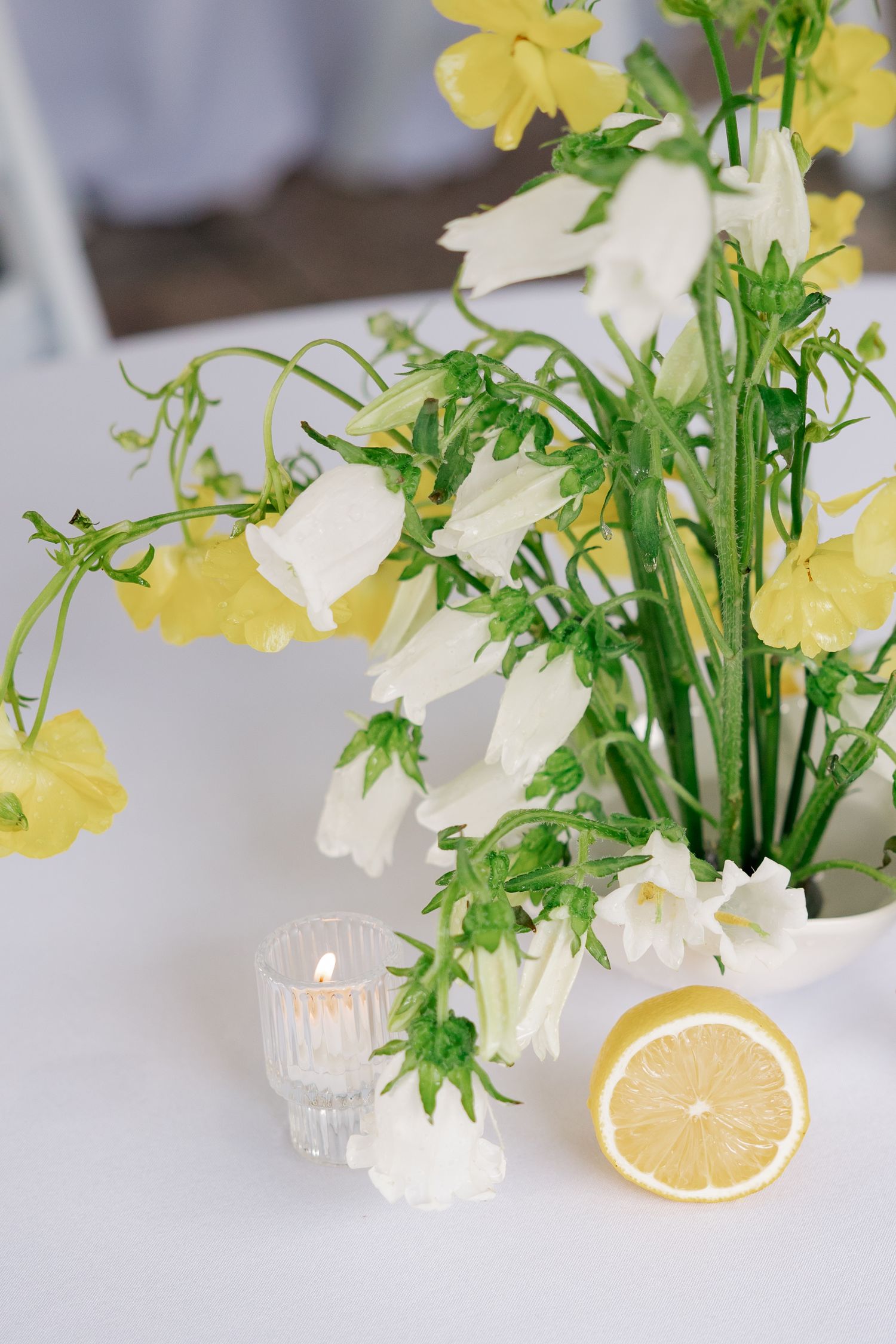 White and yellow bell flowers in a vase with a halved lemon and lit candle on a white table.