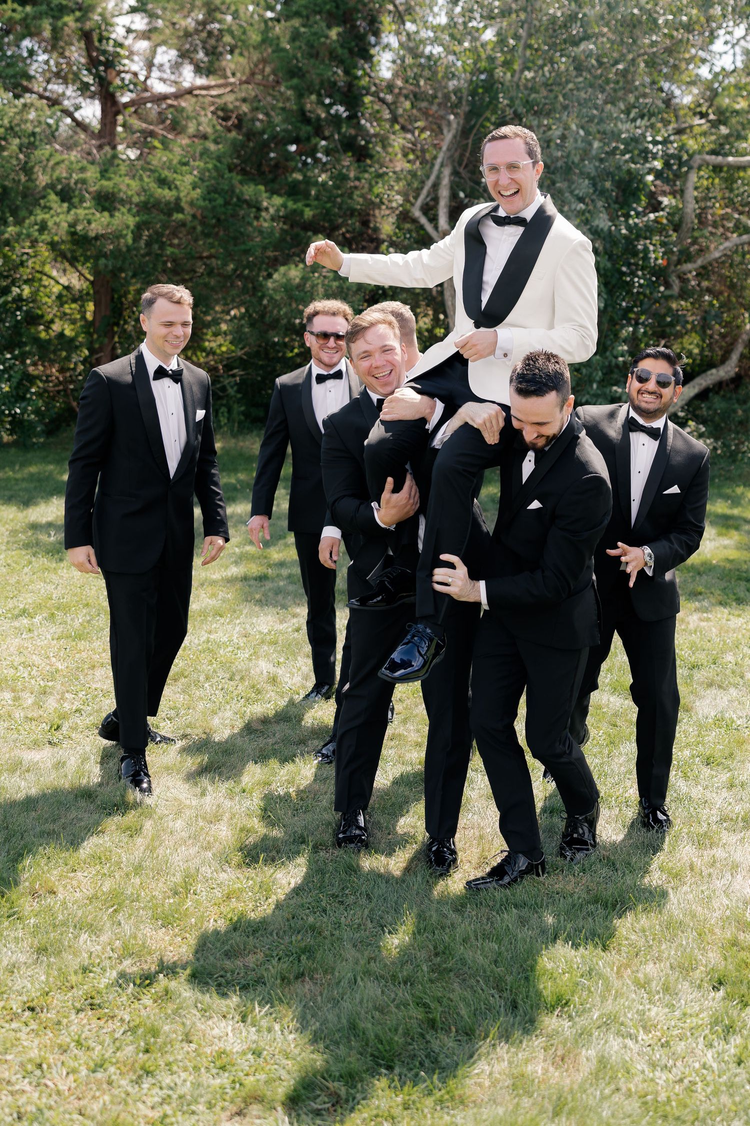 Groom in white tuxedo lifted on shoulders by groomsmen in black tuxedos on green lawn.