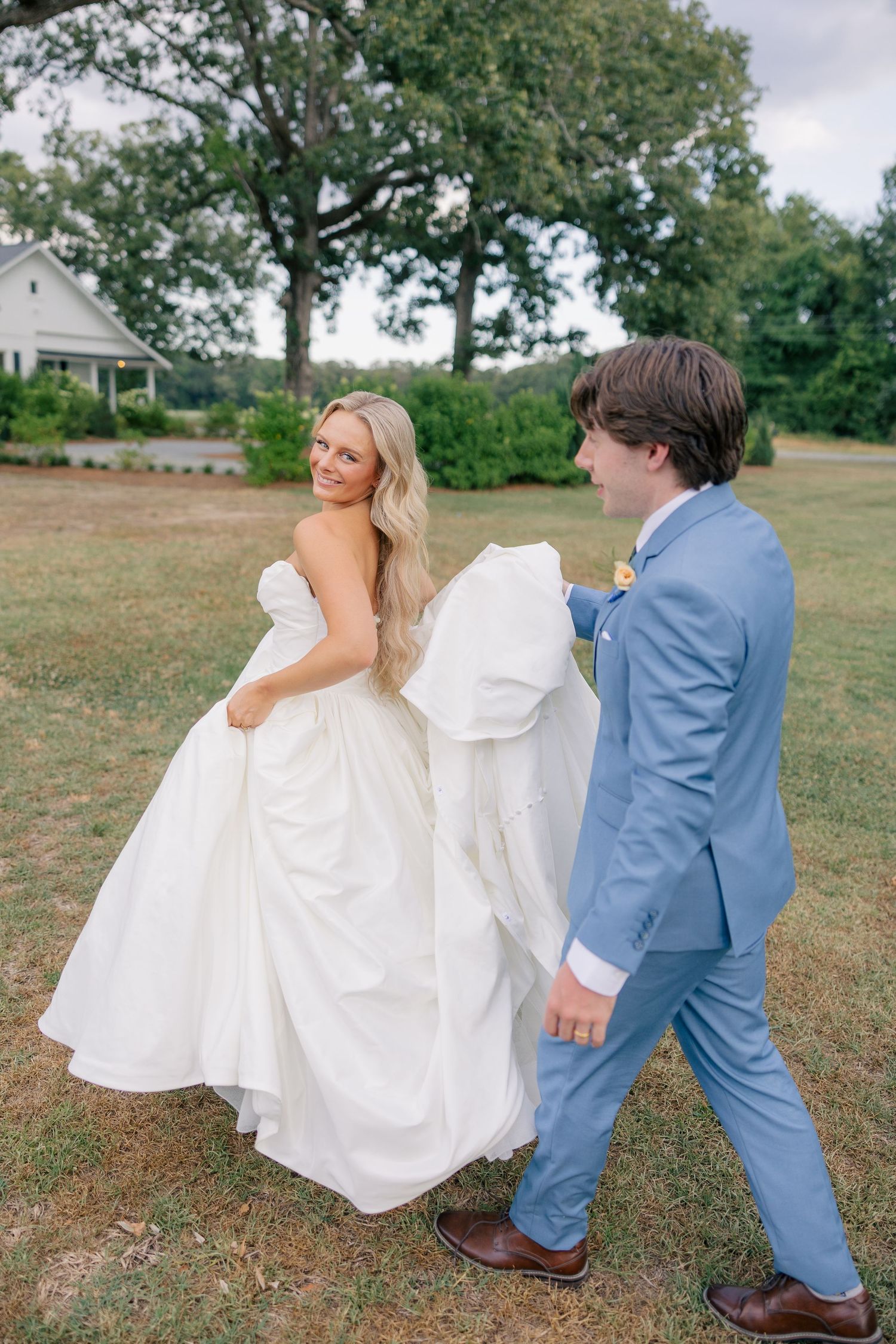 A bride in a white gown smiles as a groom in a light blue suit carries her train across a green lawn.