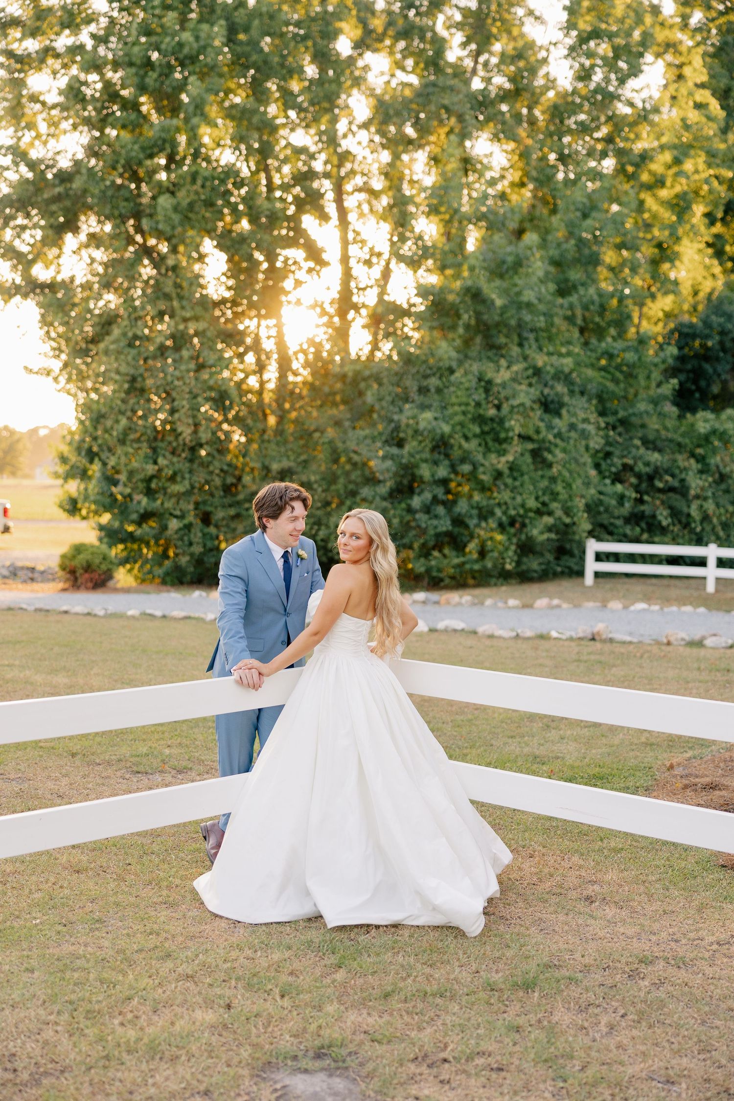 Bride and groom share romantic moment by white fence surrounded by lush green trees at sunset.