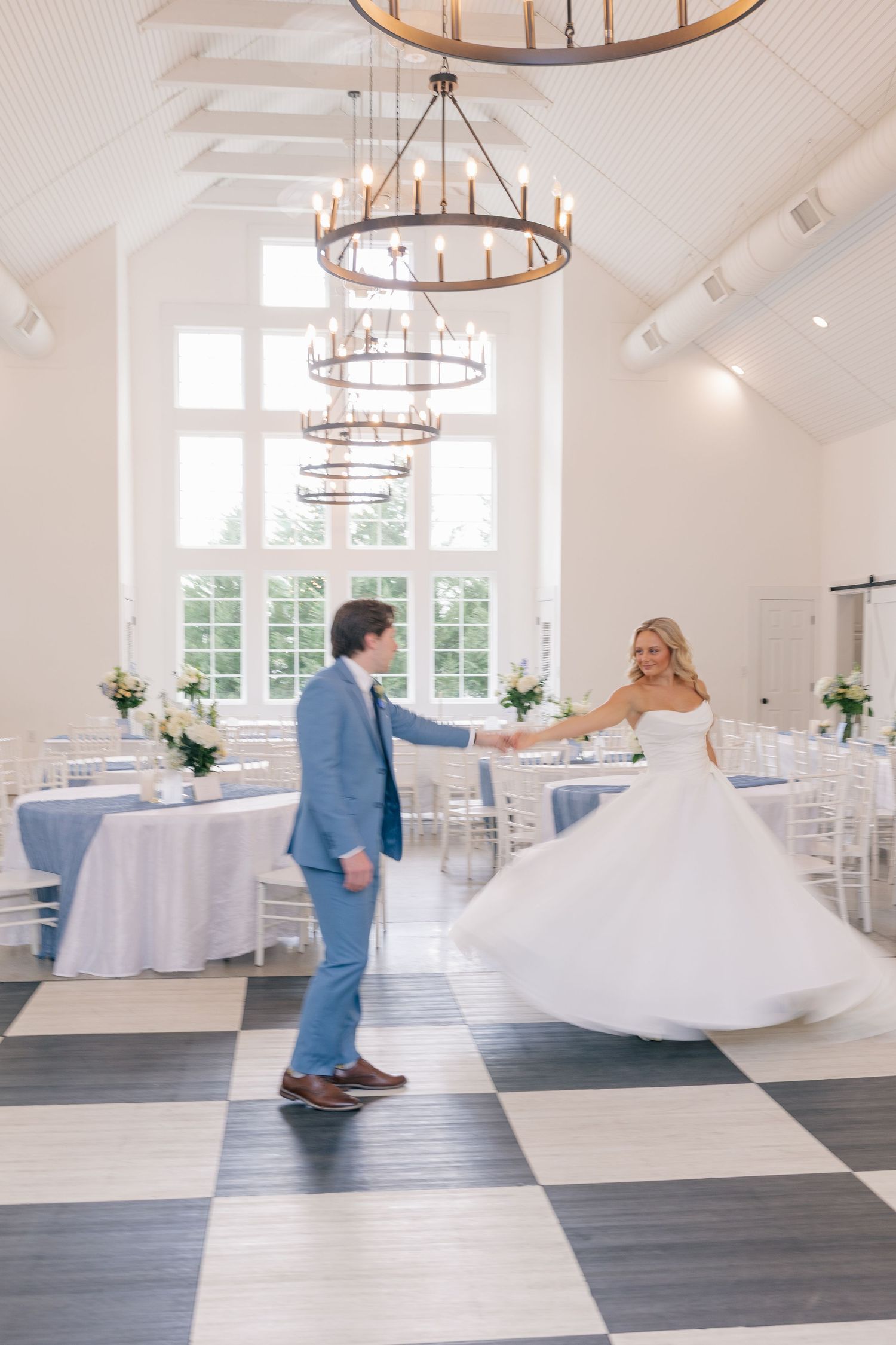 Bride and groom share first dance on checkered floor in elegant white wedding venue with chandelier.