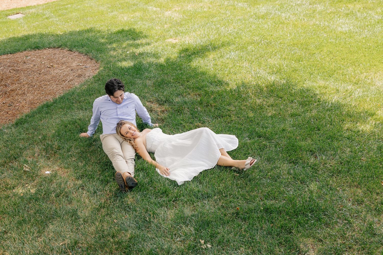 Couple in wedding attire relaxing on lush green grass, bride in white dress leaning on groom outdoors.