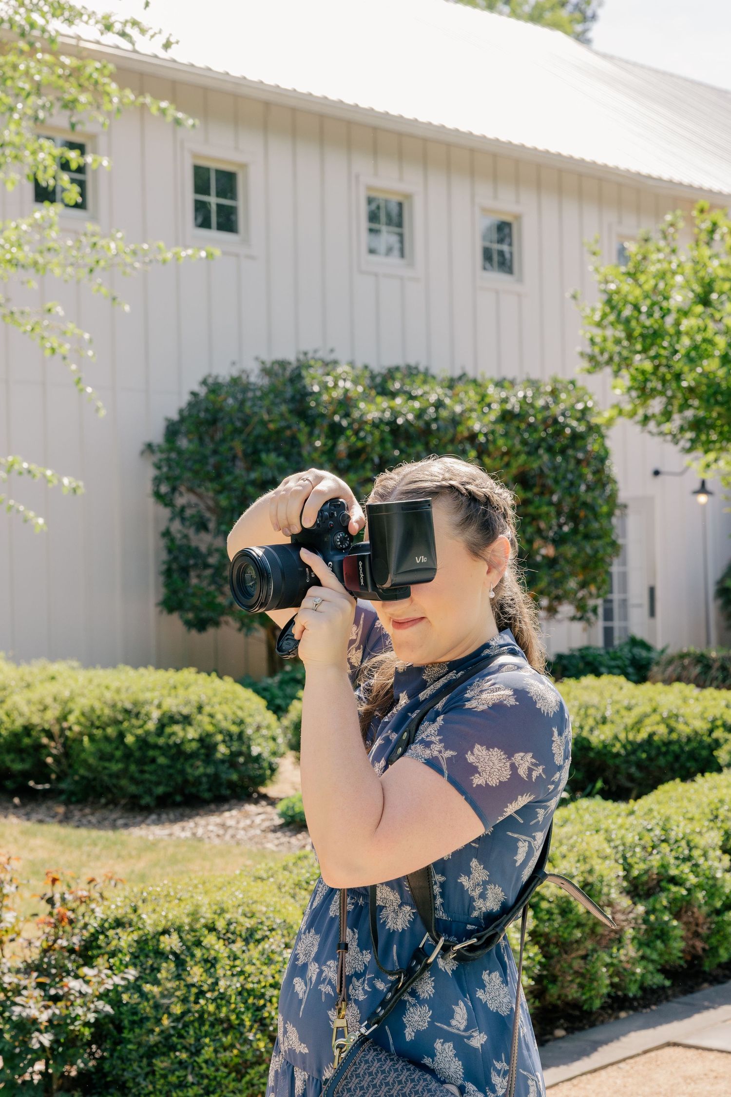 Woman in floral dress photographing with DSLR camera outdoors near white barn building surrounded by green shrubs.