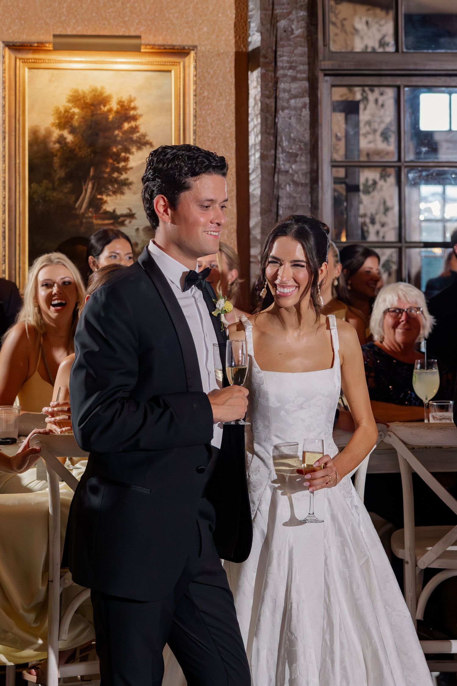 Happy bride and groom toast with champagne glasses at wedding reception surrounded by laughing guests.