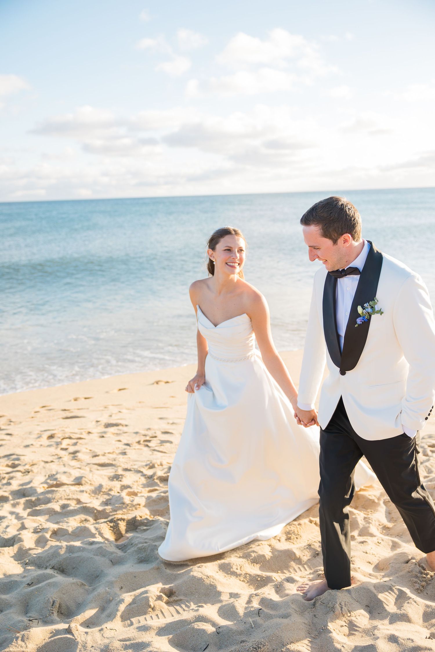 Smiling bride and groom walk hand-in-hand along a sunny beach, groom in white tuxedo jacket.