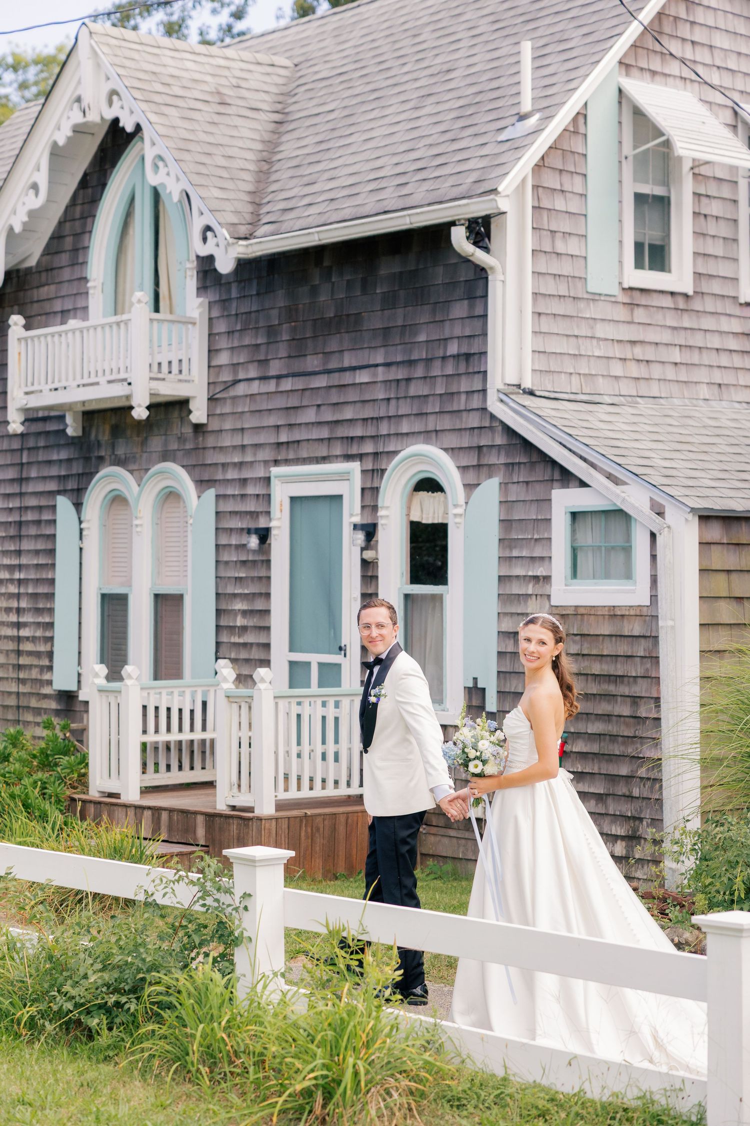 Bride and groom hold hands in front of a charming gray shingle Victorian cottage with white fence.