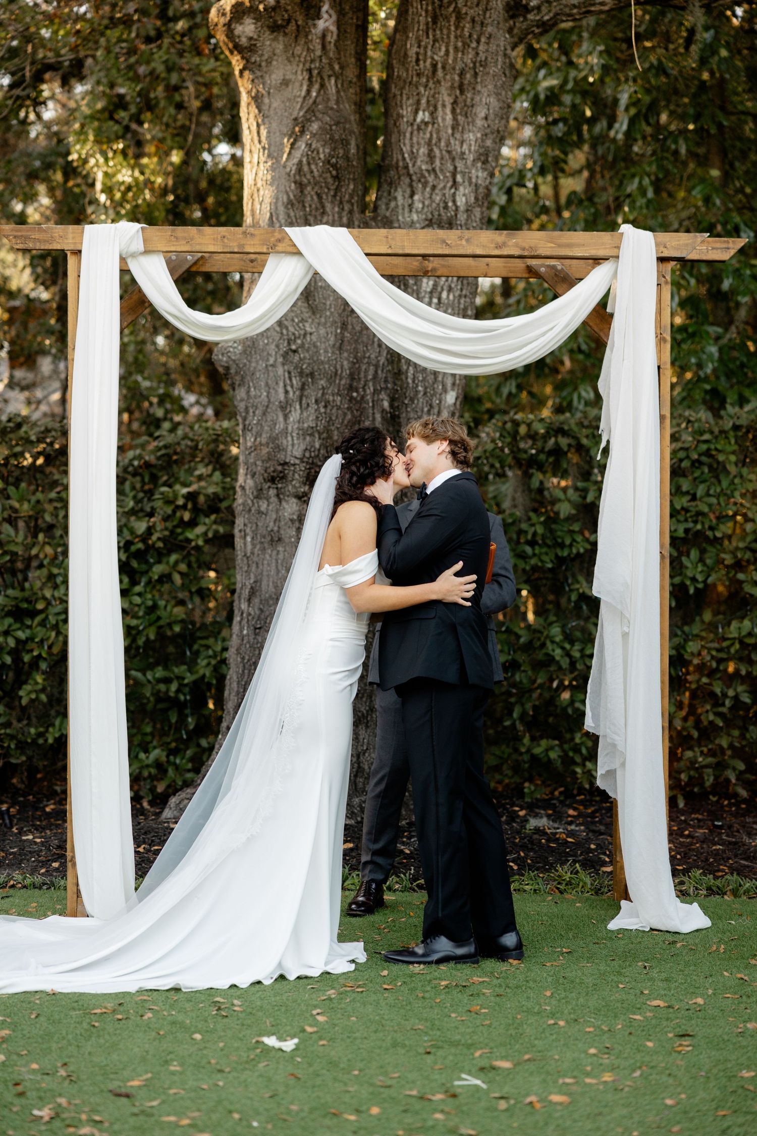 A wedding ceremony takes place under a simple white draped arch against a backdrop of mature trees.