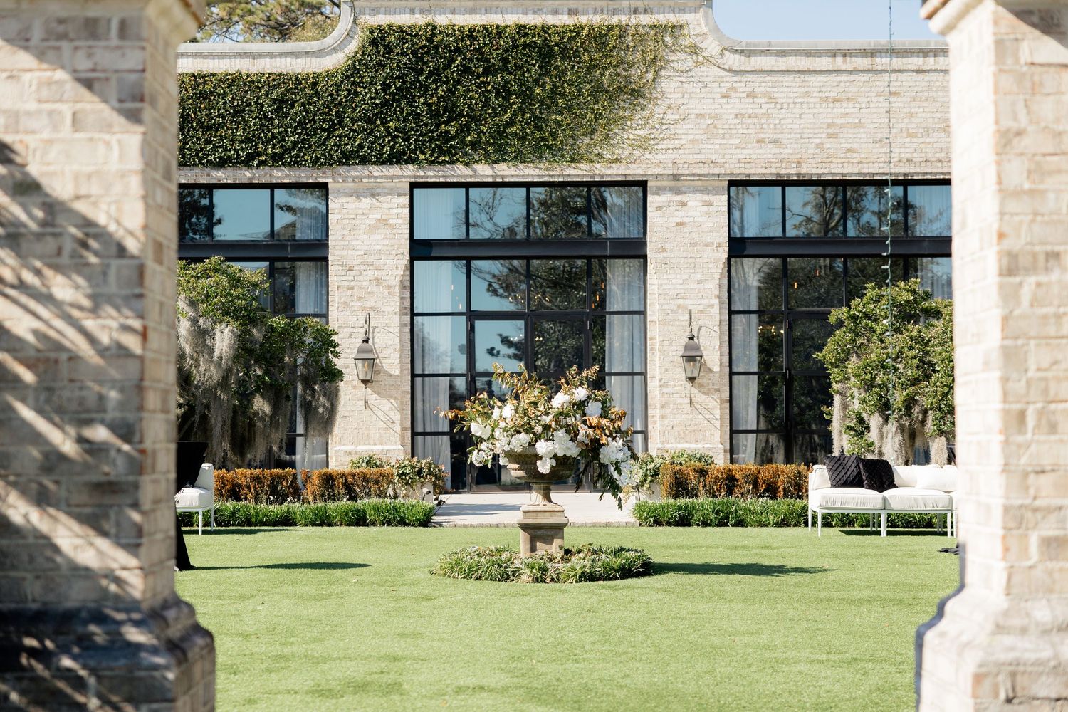 Elegant outdoor wedding venue with stone architecture, large windows, manicured lawn and floral arrangements.