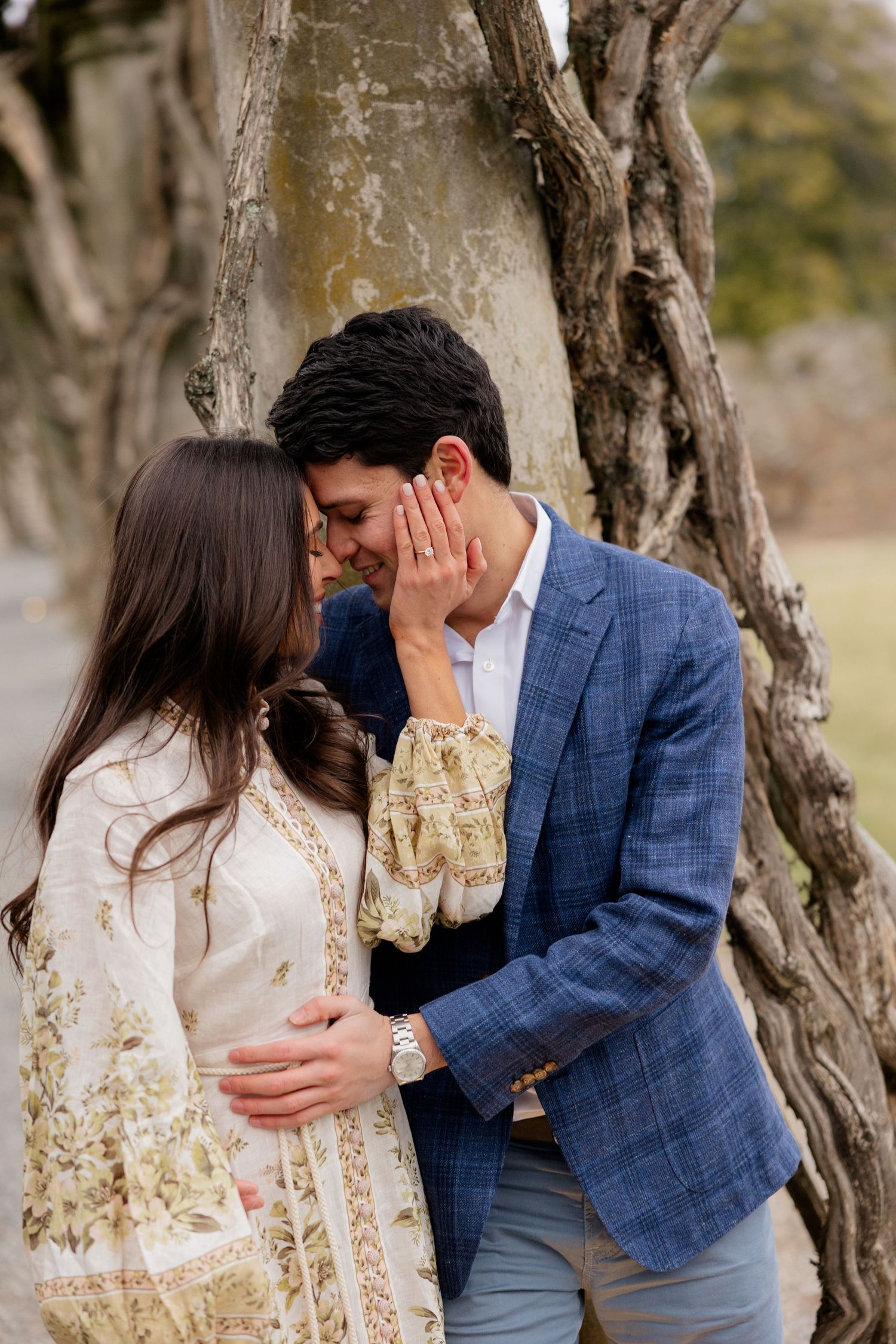 A romantic couple in a floral dress and blue blazer share an intimate moment beside a tree trunk in a park setting.