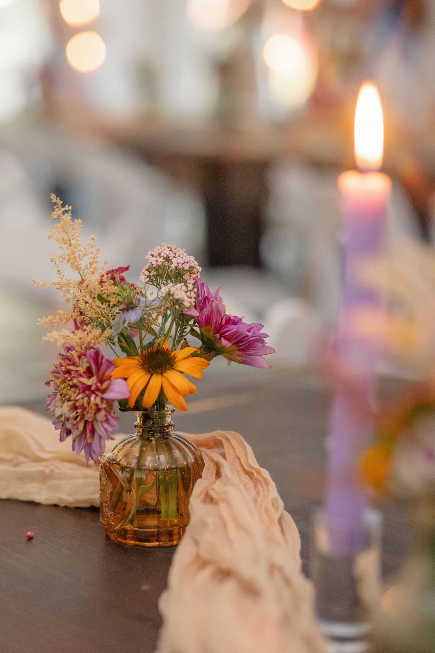 Colorful wildflower bouquet in amber glass vase with lit candle on wedding reception table.