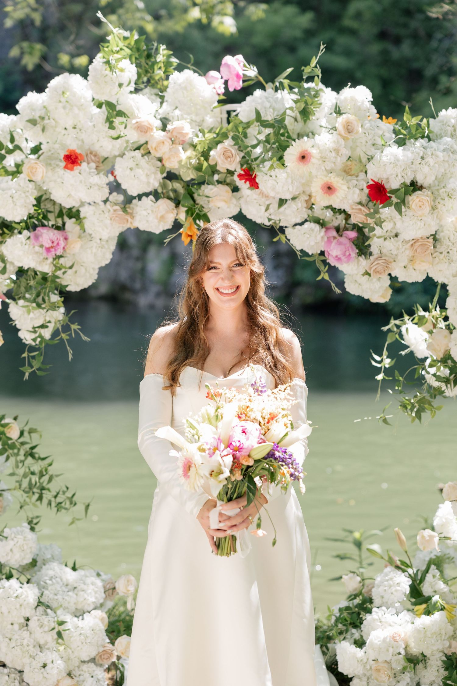 Radiant bride holding a pastel bouquet standing beneath a lush white floral wedding arch.
