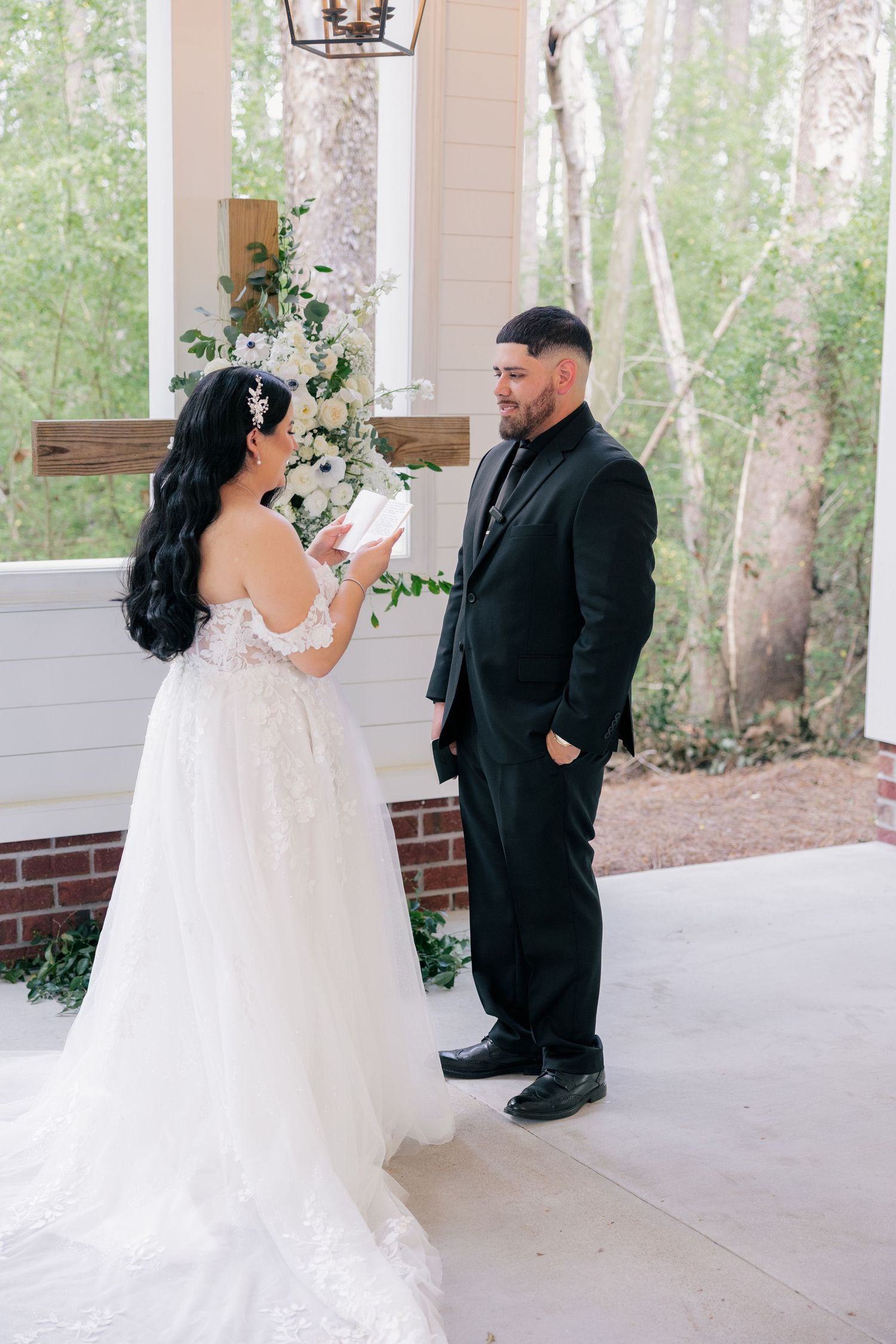 Couple exchanging vows on a covered porch with floral decorations and birch trees in the background.