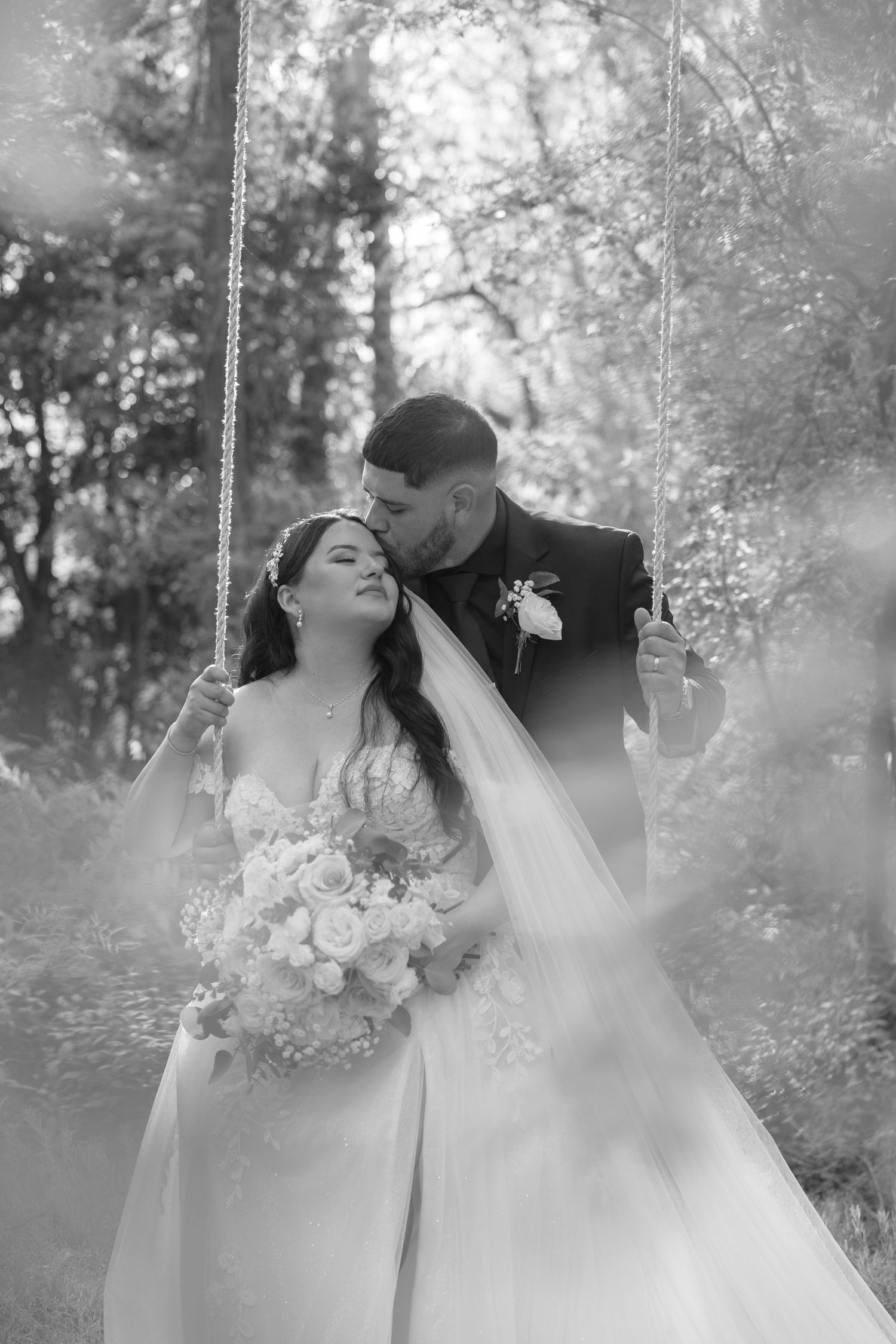 A black and white romantic wedding portrait of a couple on a swing surrounded by dreamy foliage.