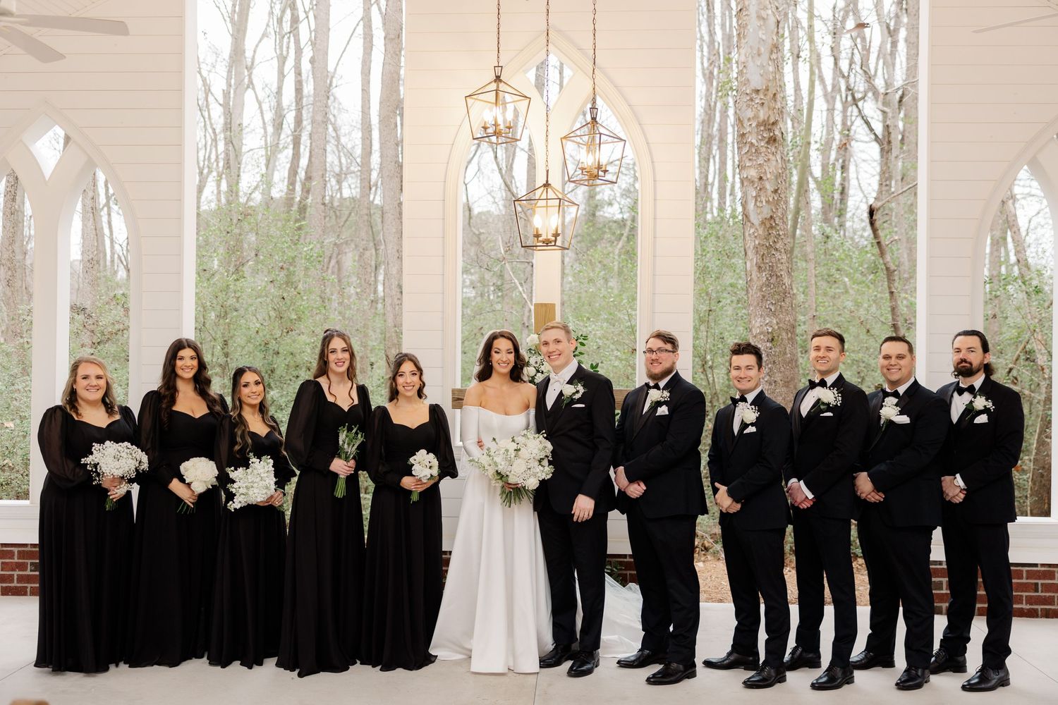 A wedding party in black and white attire poses in formation inside a bright white chapel.